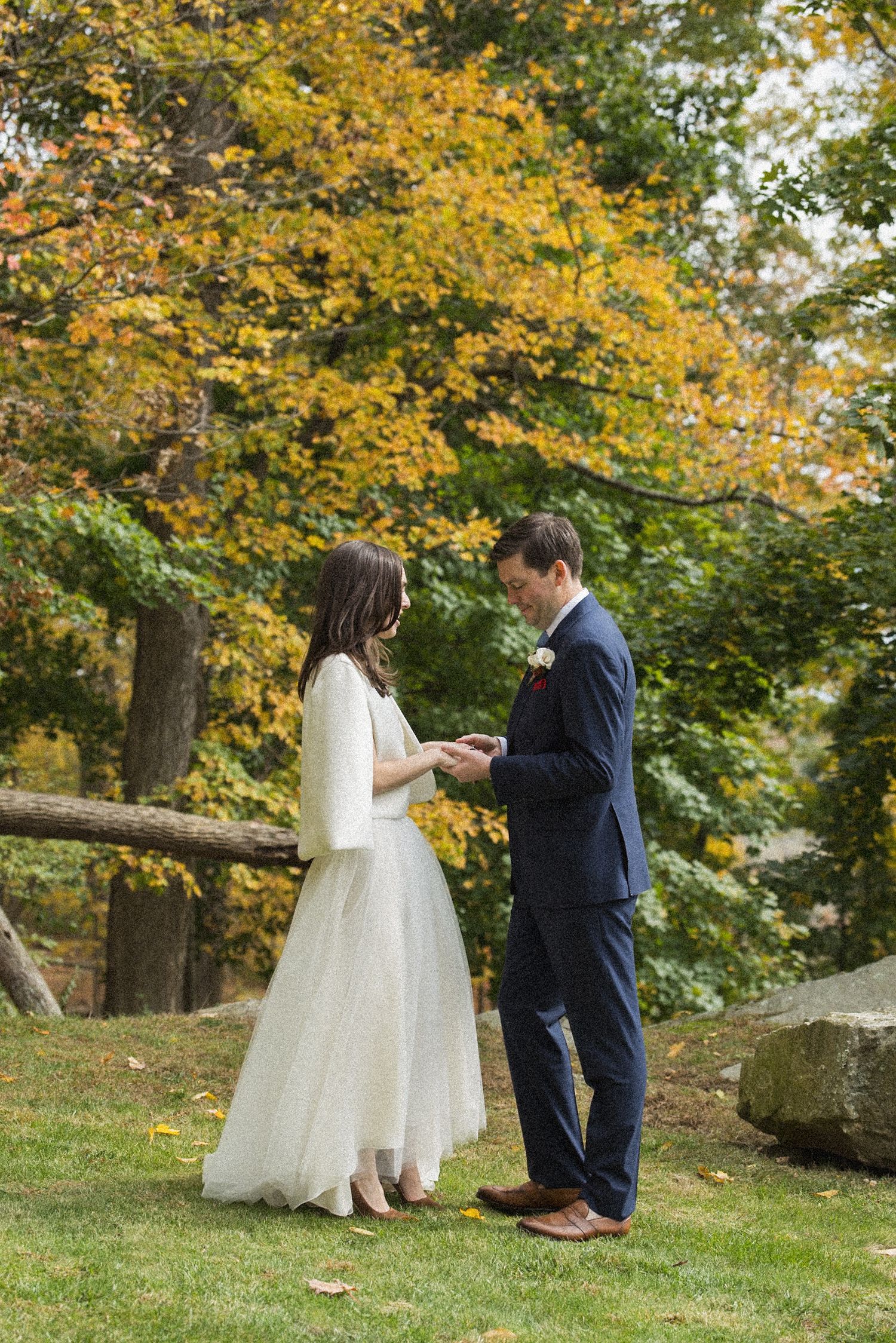 A bride in a white tulle skirt and a groom in a navy suit hold hands during an outdoor autumn wedding ceremony beneath golden trees.