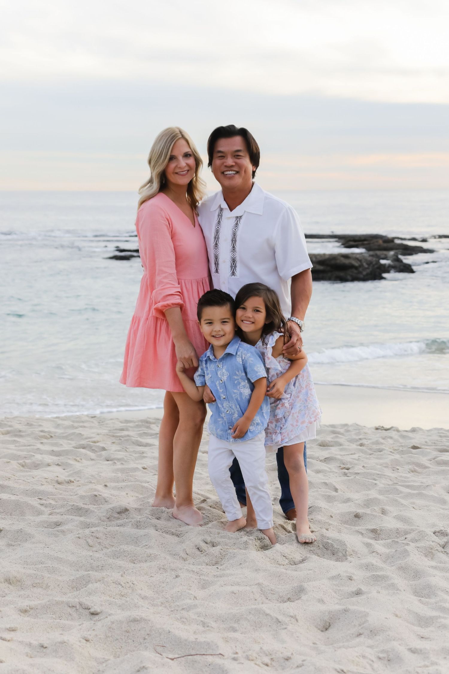 Family wearing coordinated white and blue outfits posing for photos on a beach at dusk.