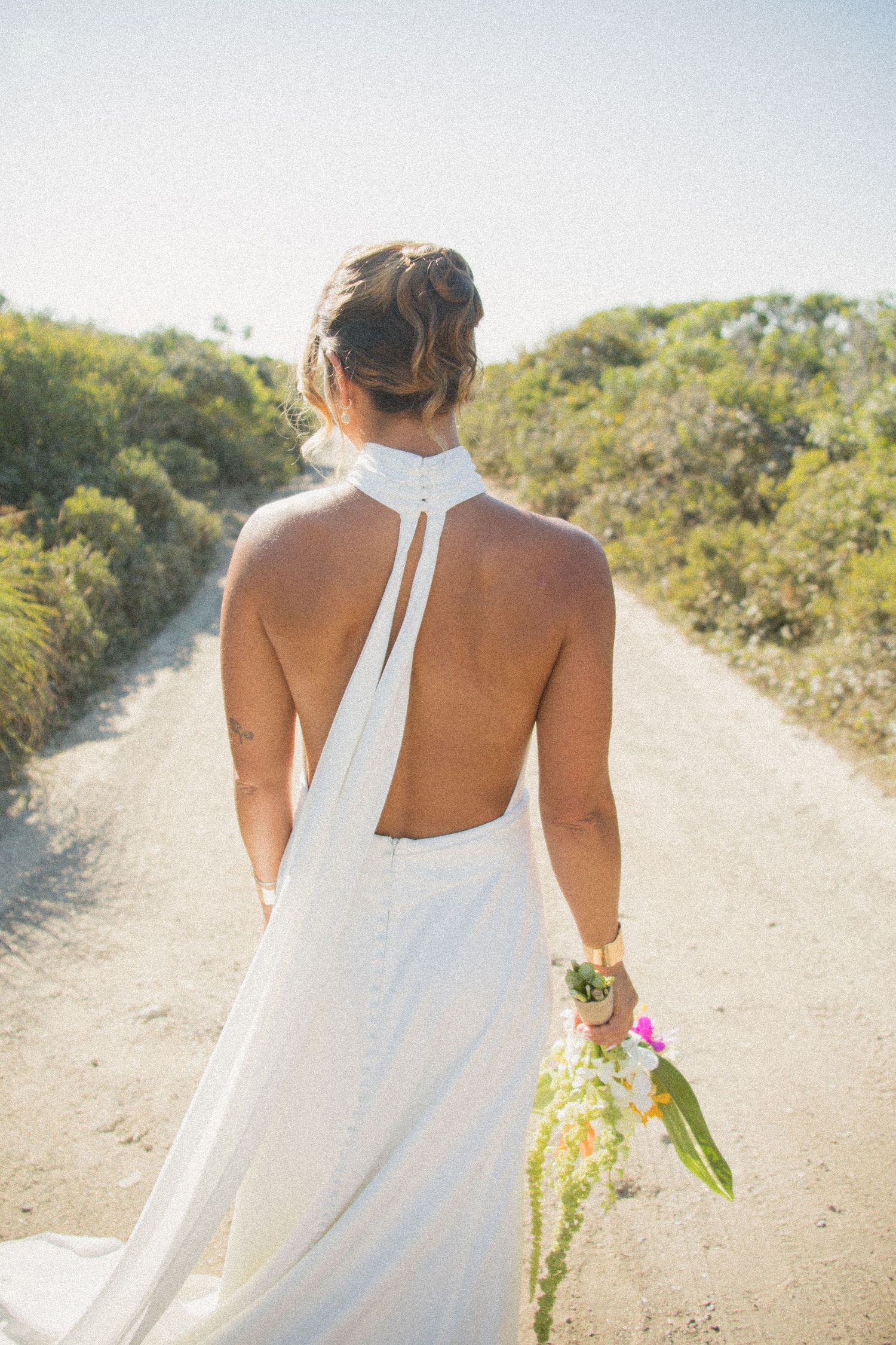 Back view of bride in halter wedding dress with open back detail walking on dirt path holding bouquet.
