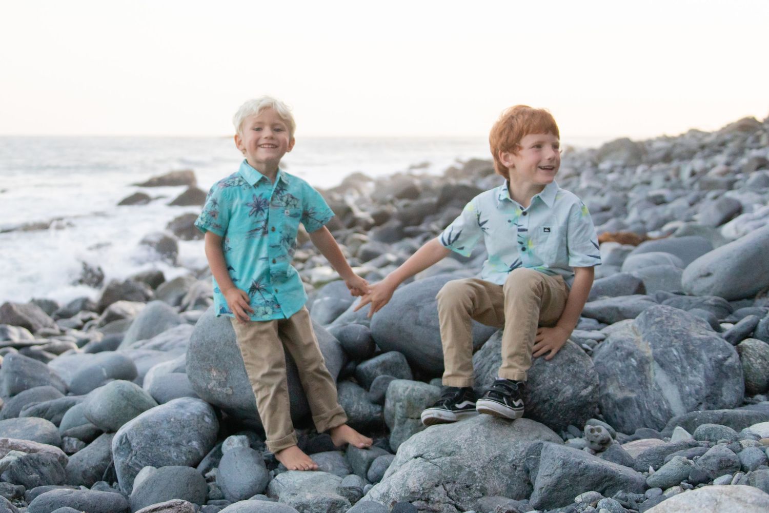 Children sitting on large rocks at a beach with cliffs in background.
