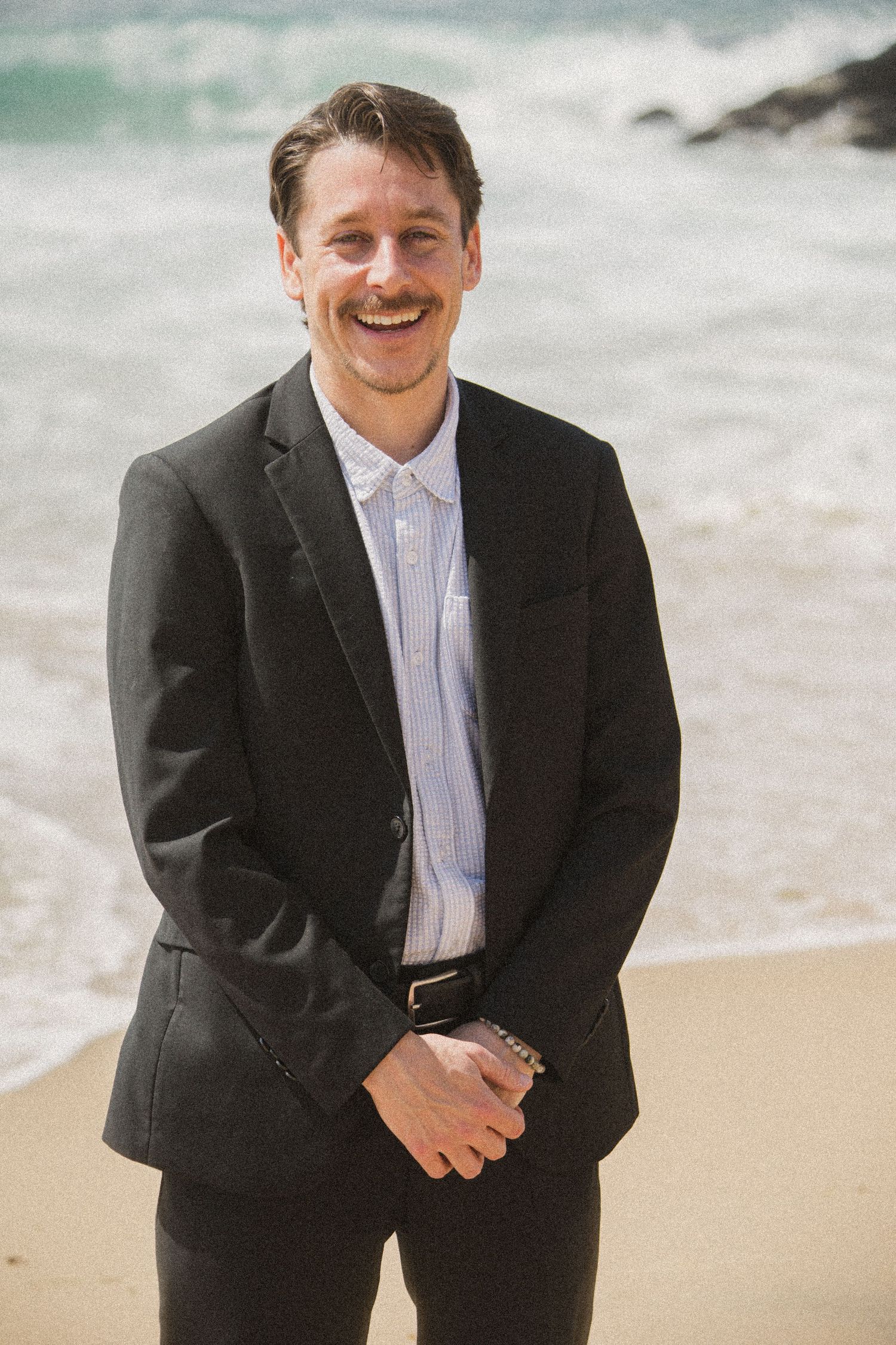 Groom laughs joyfully during beach wedding photoshoot wearing classic black suit.
