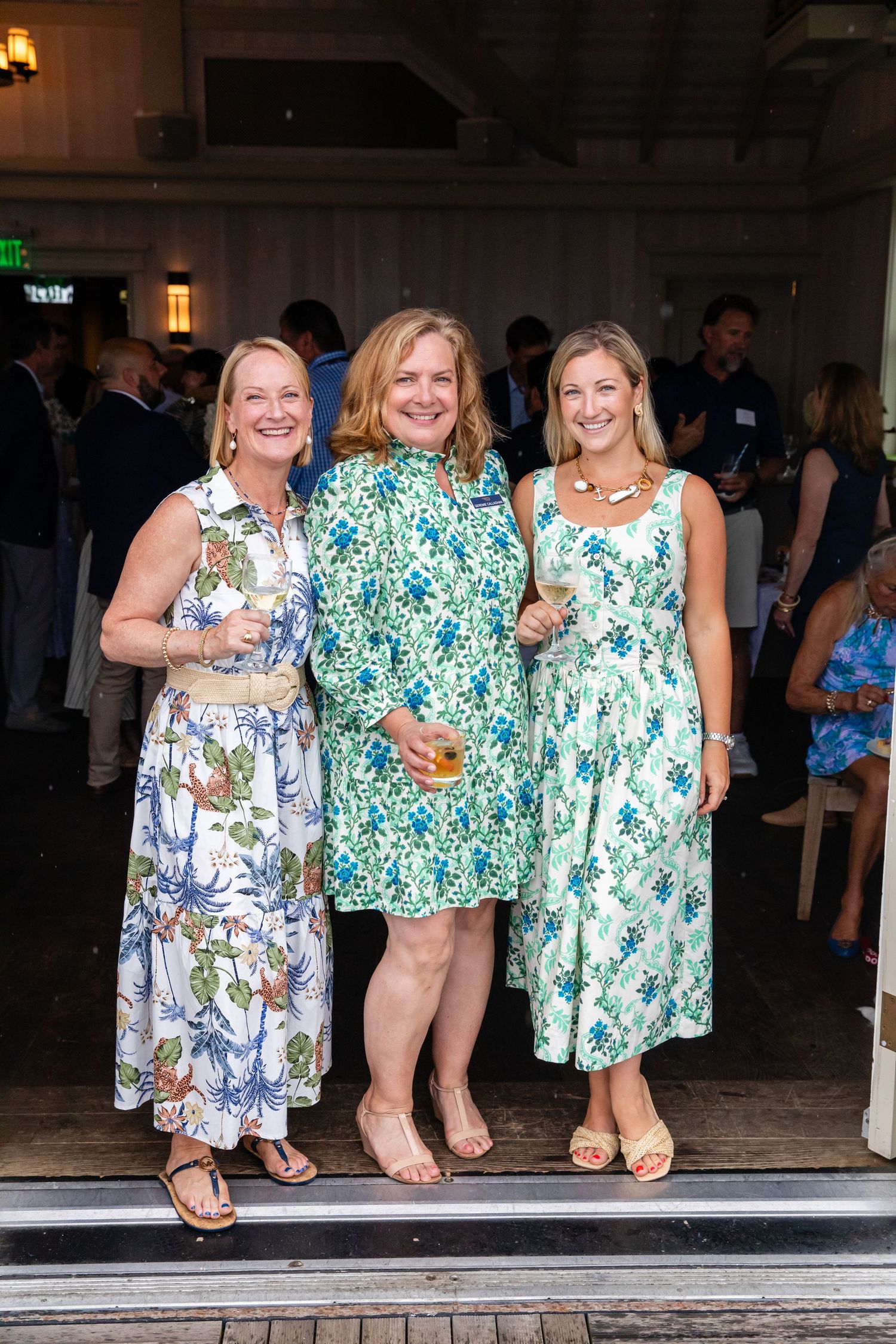 Three women in floral dresses smile together at an outdoor summer social event.