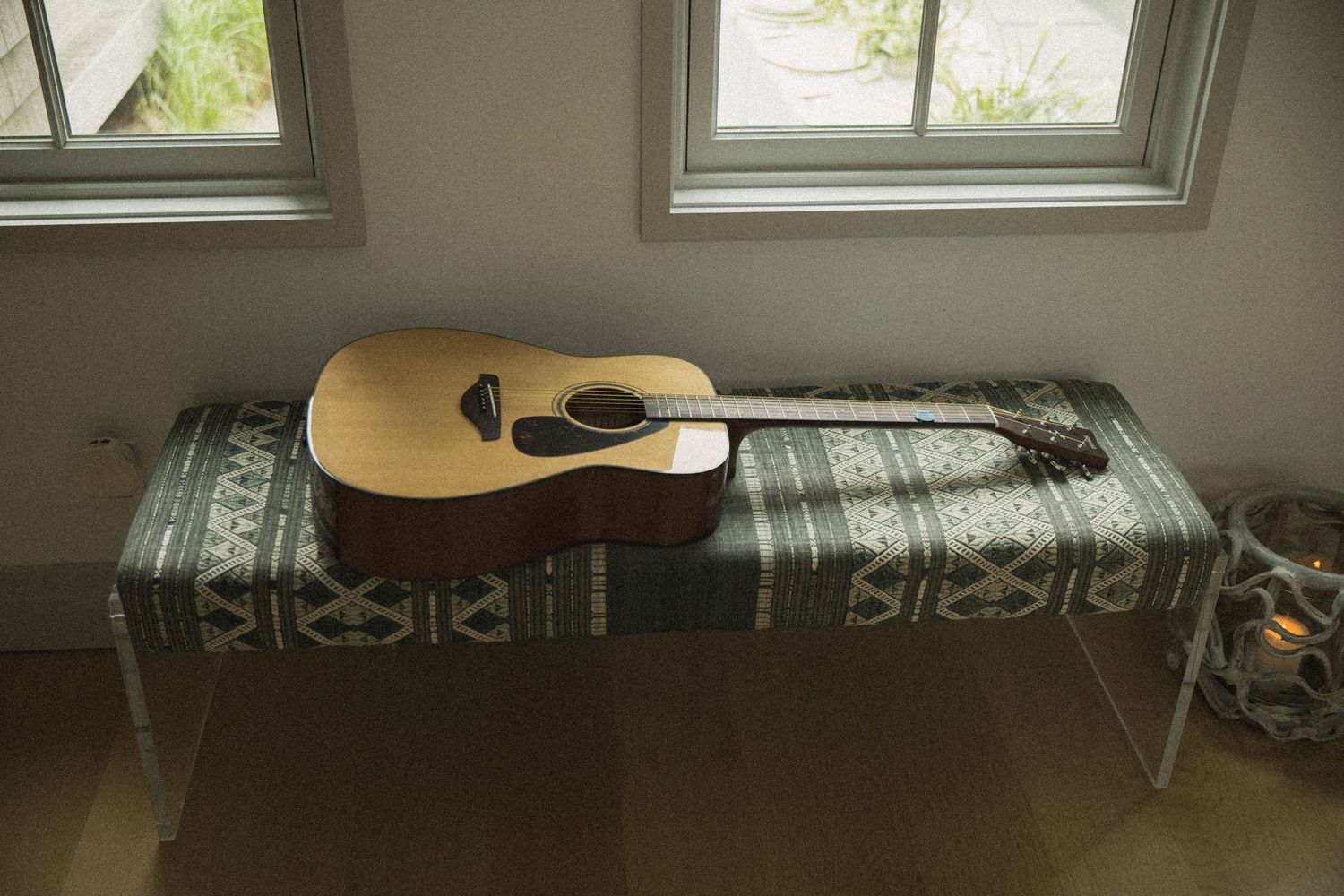 Acoustic guitar resting on a table with a patterned cloth cover near sunlit windows in a bright room.