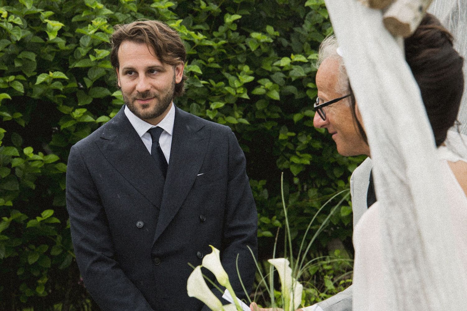 Groom in dark suit smiling at bride during outdoor wedding ceremony with lush green foliage backdrop.