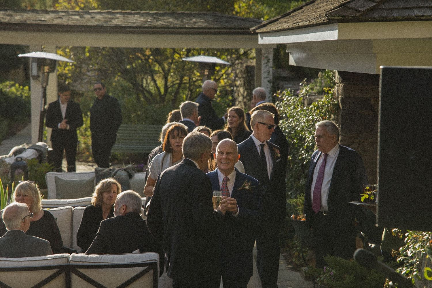 Guests in formal attire mingle and chat on an outdoor patio at a garden reception, with seating areas and greenery in the background.