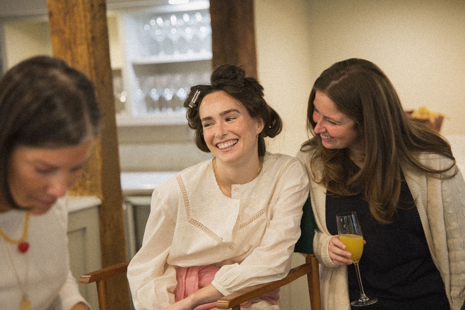 Two smiling women laugh together at a table while one holds a glass of orange juice in a bright, cozy room.