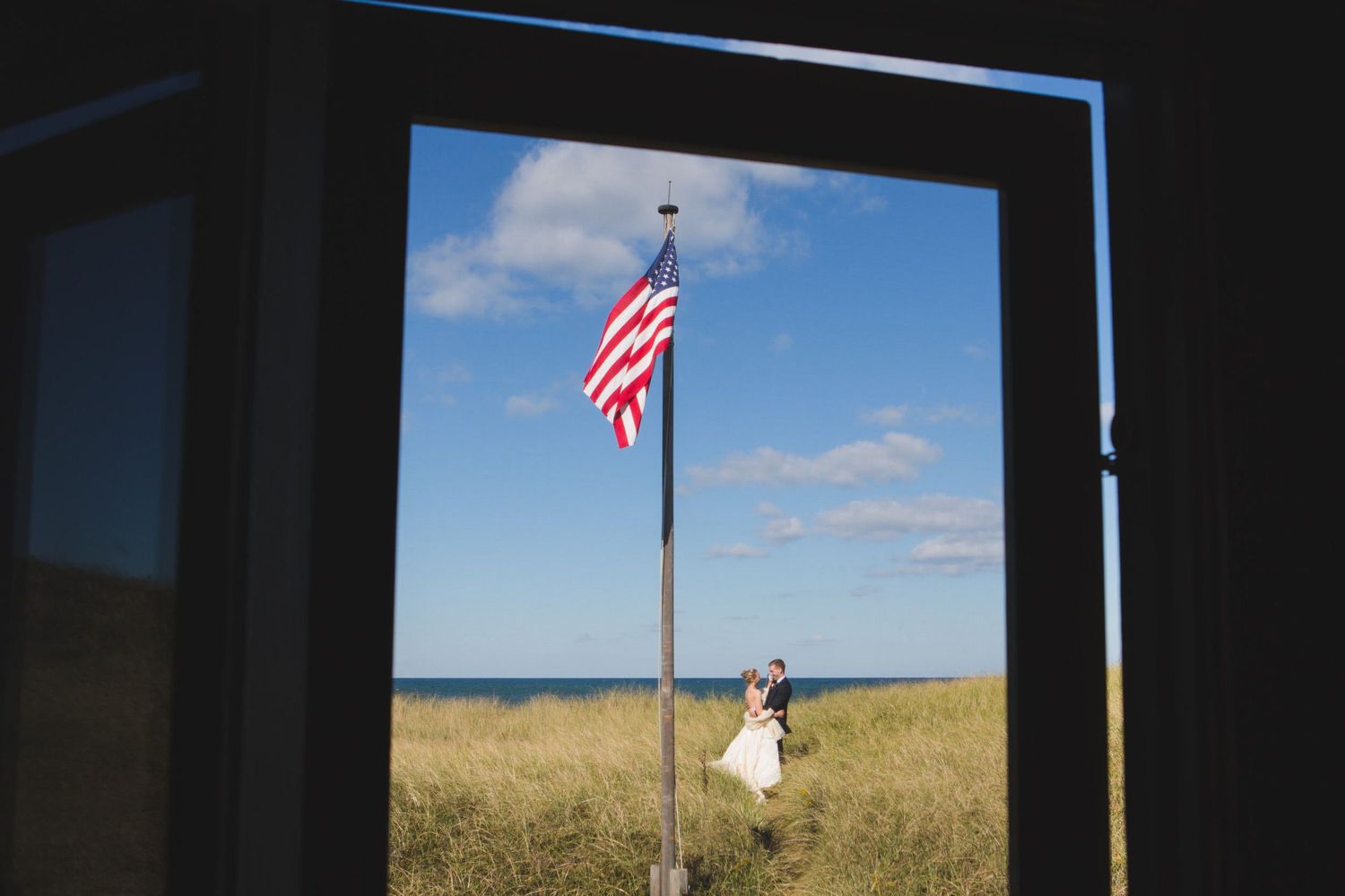 American flag waves against blue sky as person walks through golden wheat field on sunny summer day.