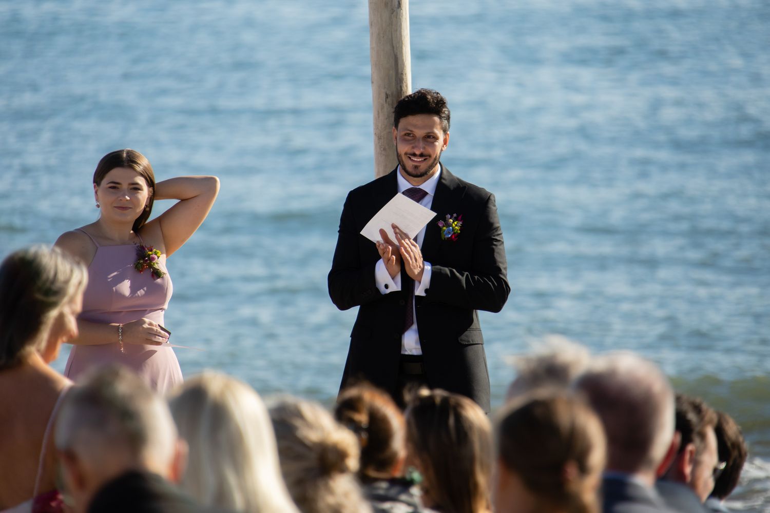 An outdoor beach wedding ceremony with attendees watching the proceedings by the ocean.