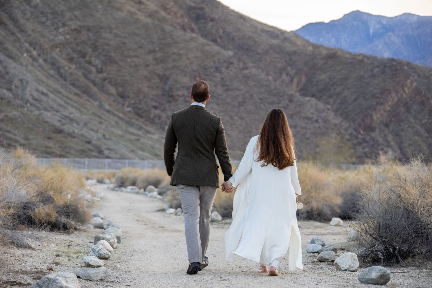 A couple walks along a desert path with mountains in the background during sunset.