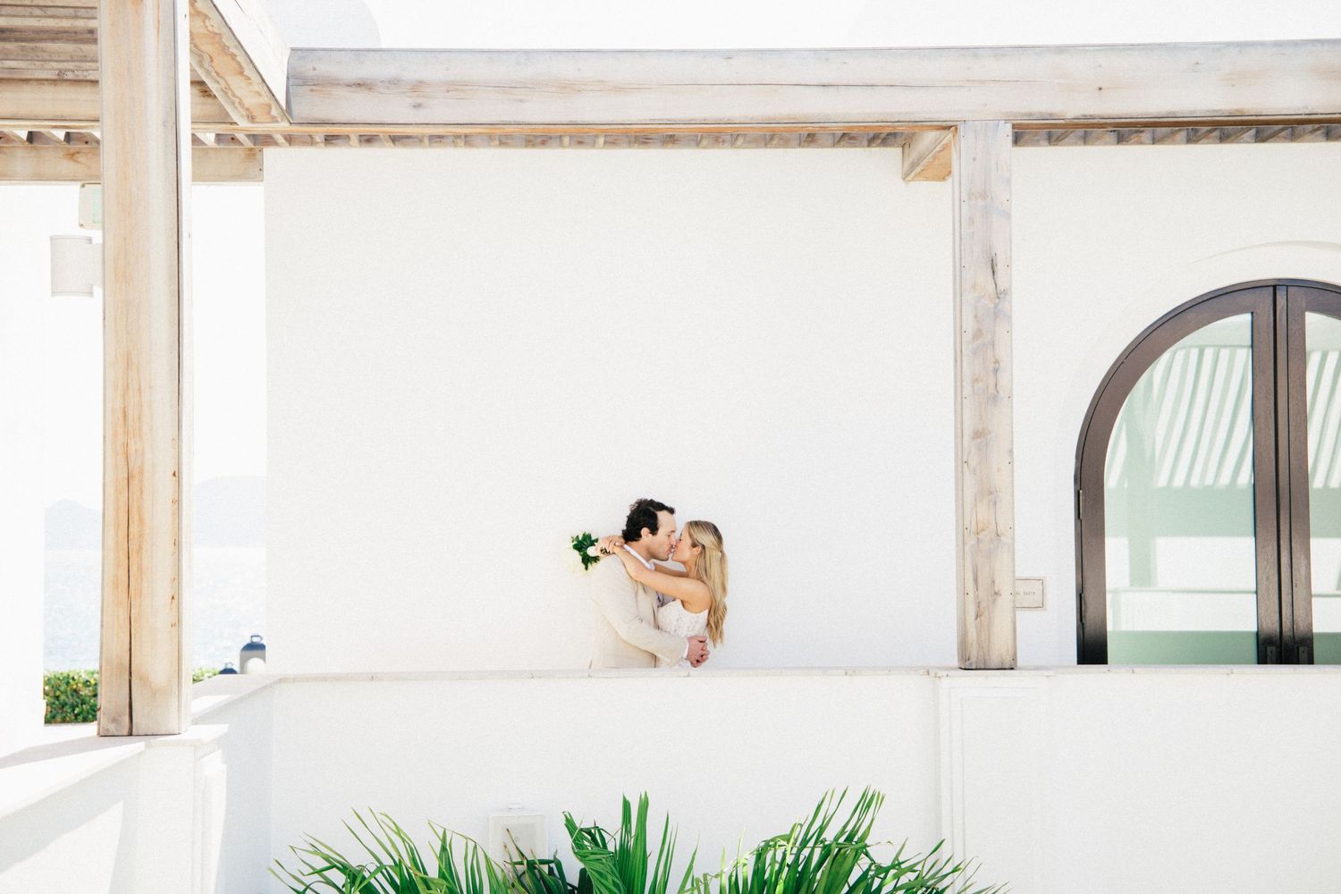 A minimalist white balcony wedding photo with architectural arches and tropical plants below.