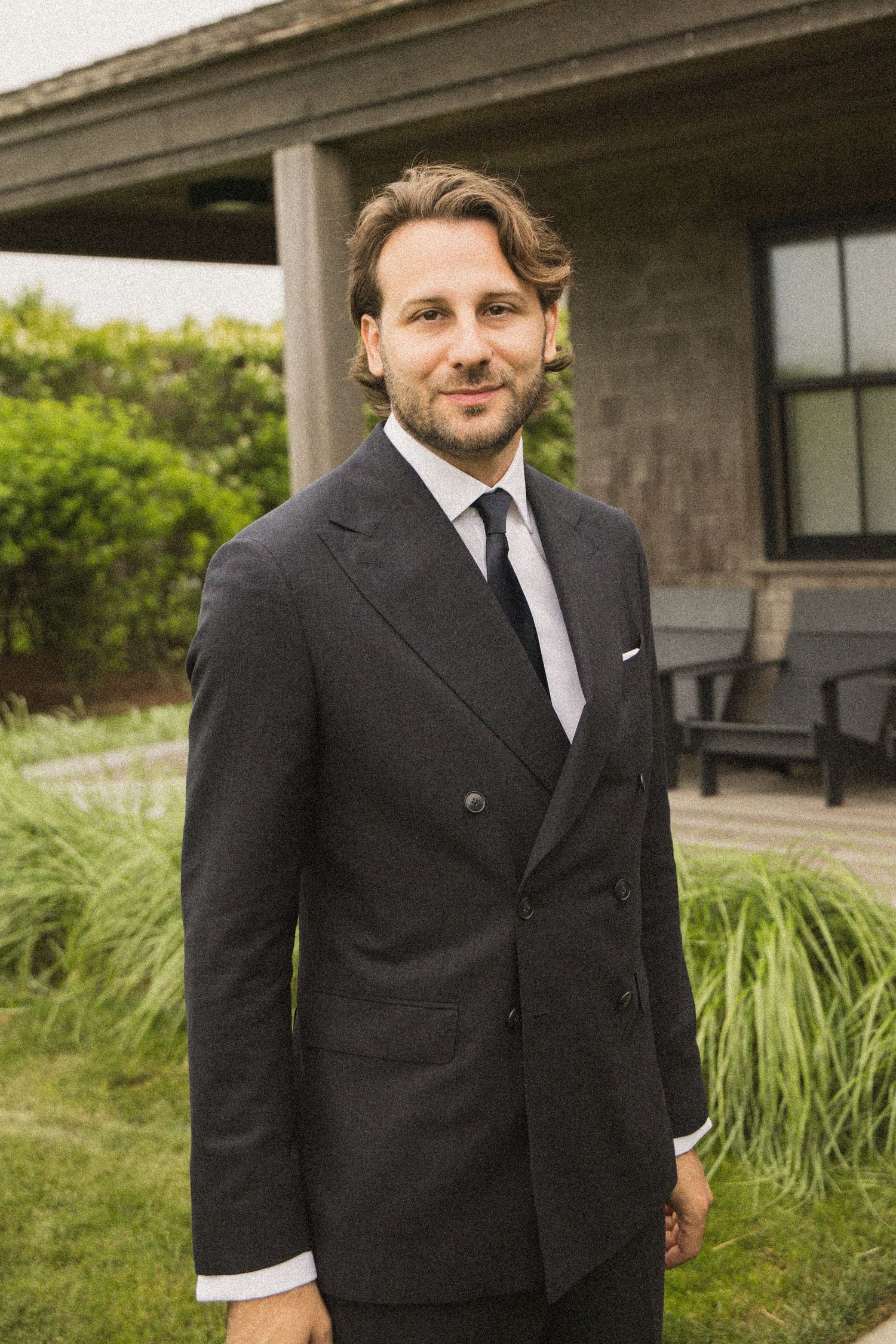 Man in dark double-breasted suit and tie standing outdoors near a building with green landscaping.