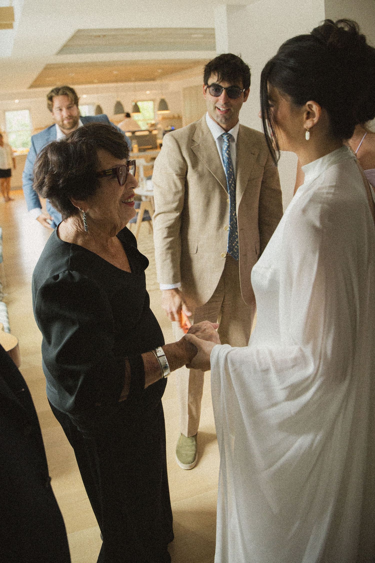 Elderly woman in black shakes hands with bride in white dress, man in beige suit watches indoors.