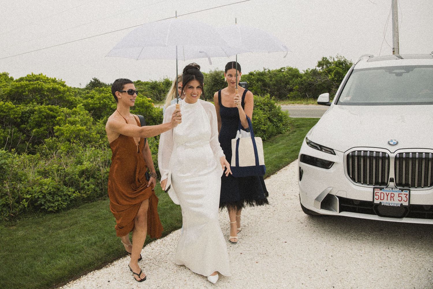Bride in white gown walks with two guests near a BMW SUV, one holding a clear umbrella on a cloudy day.