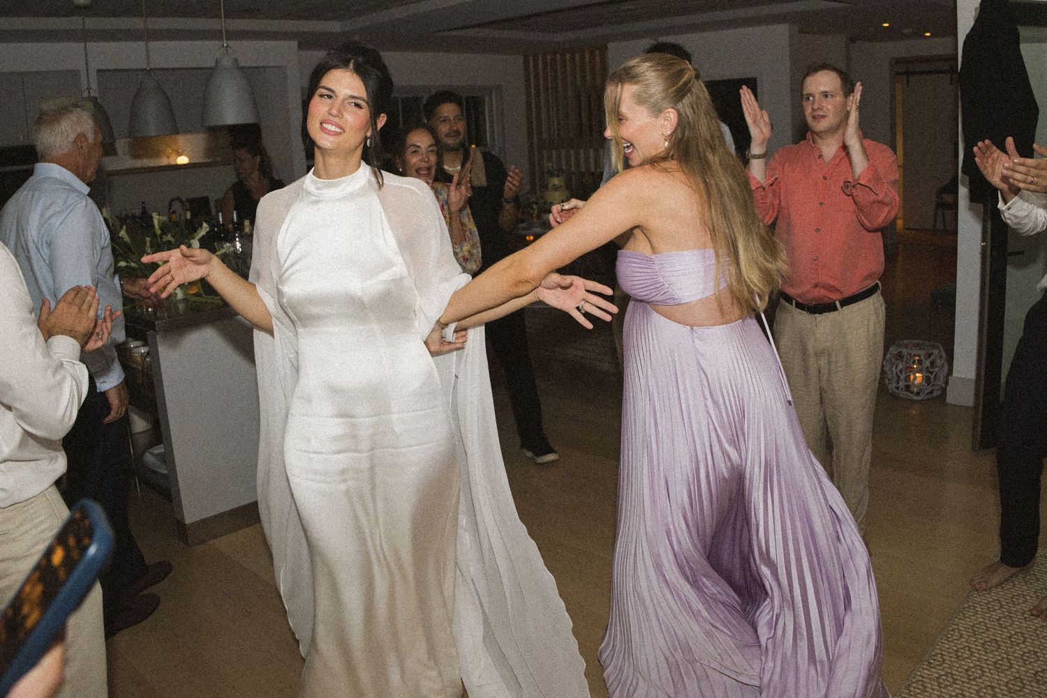Two women dancing elegantly at a wedding reception, one in white bridal attire and one in a lavender gown.