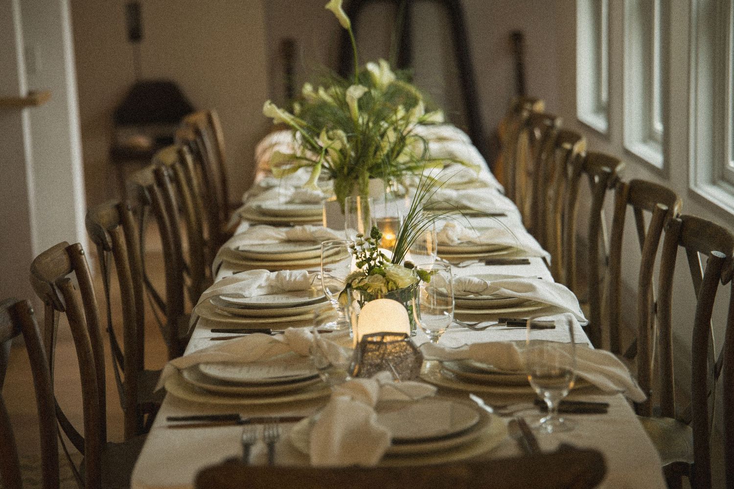Elegantly set long dining table with white linens, gold-rimmed plates, crystal glasses, and fresh floral centerpieces in a warmly lit room.