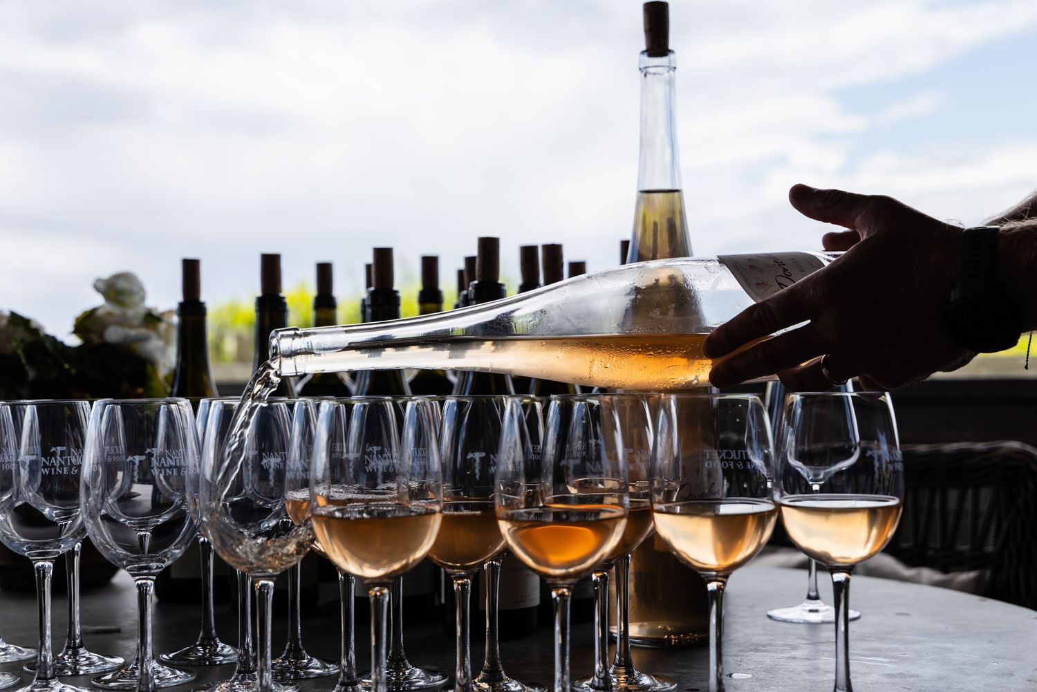 Person pouring rose wine into elegant glasses at an outdoor wine tasting event with bottles in background.