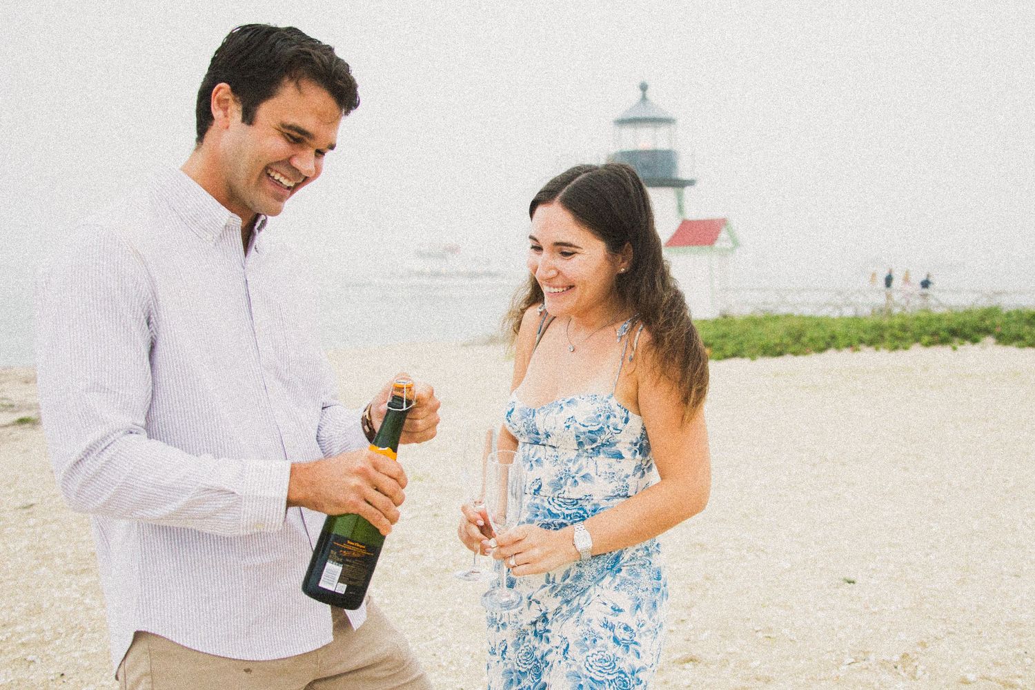 Couple celebrating on beach, man opening champagne bottle, woman in floral dress, lighthouse in background.
