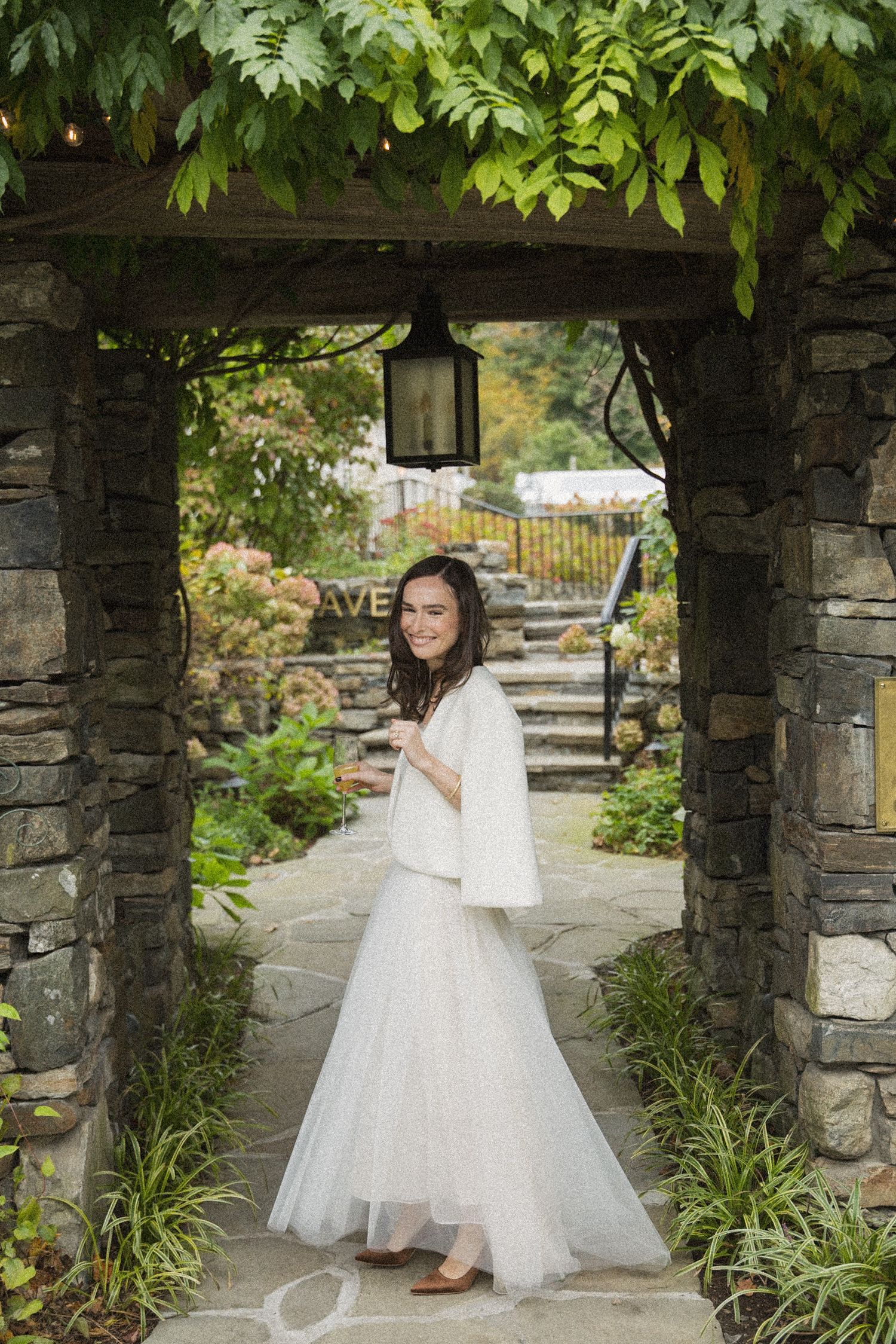 Bride in a white tulle skirt and cream shrug smiles over her shoulder beneath a stone archway with a hanging lantern and ivy.