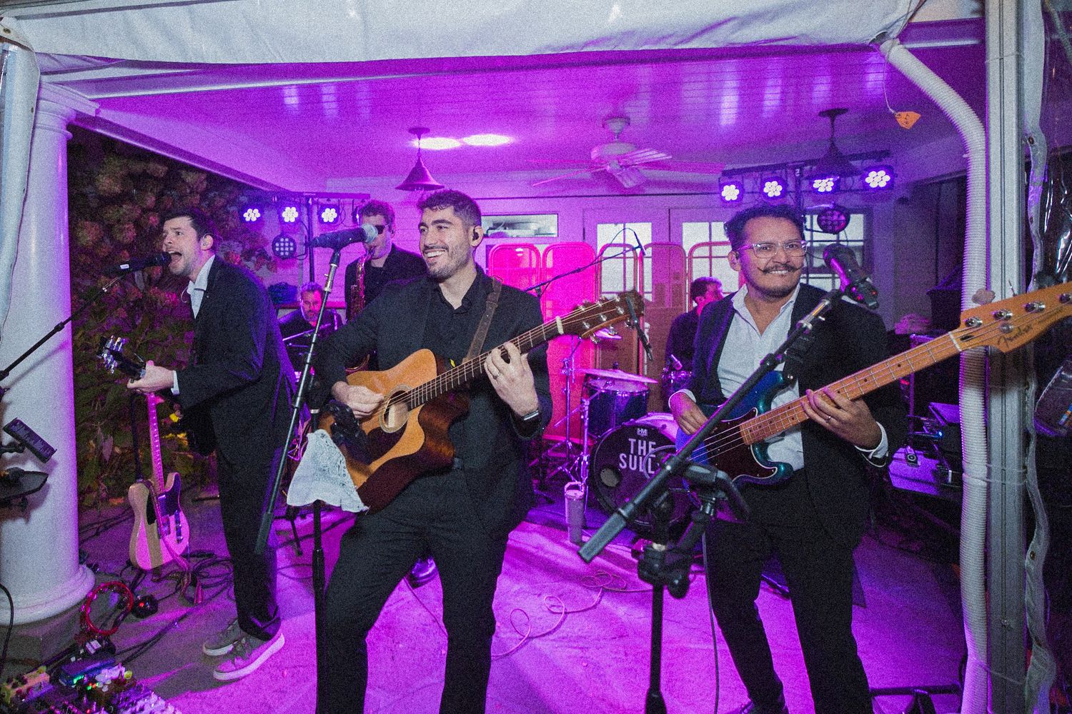 Live band members in dark outfits play guitars on a stage lit by purple lighting at a wedding reception.