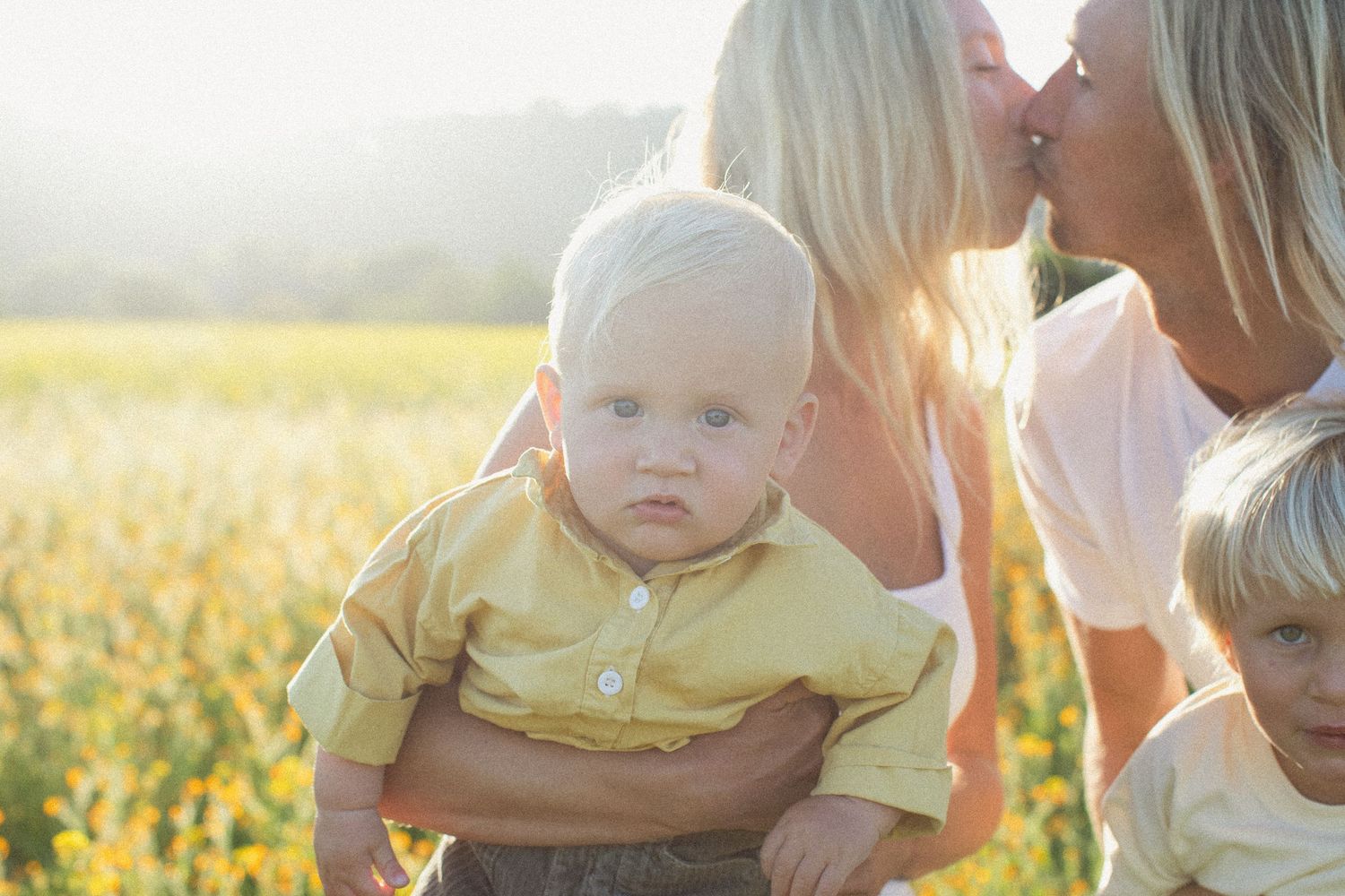 A baby in a yellow shirt sits outdoors in a sunlit field of wildflowers during golden hour.