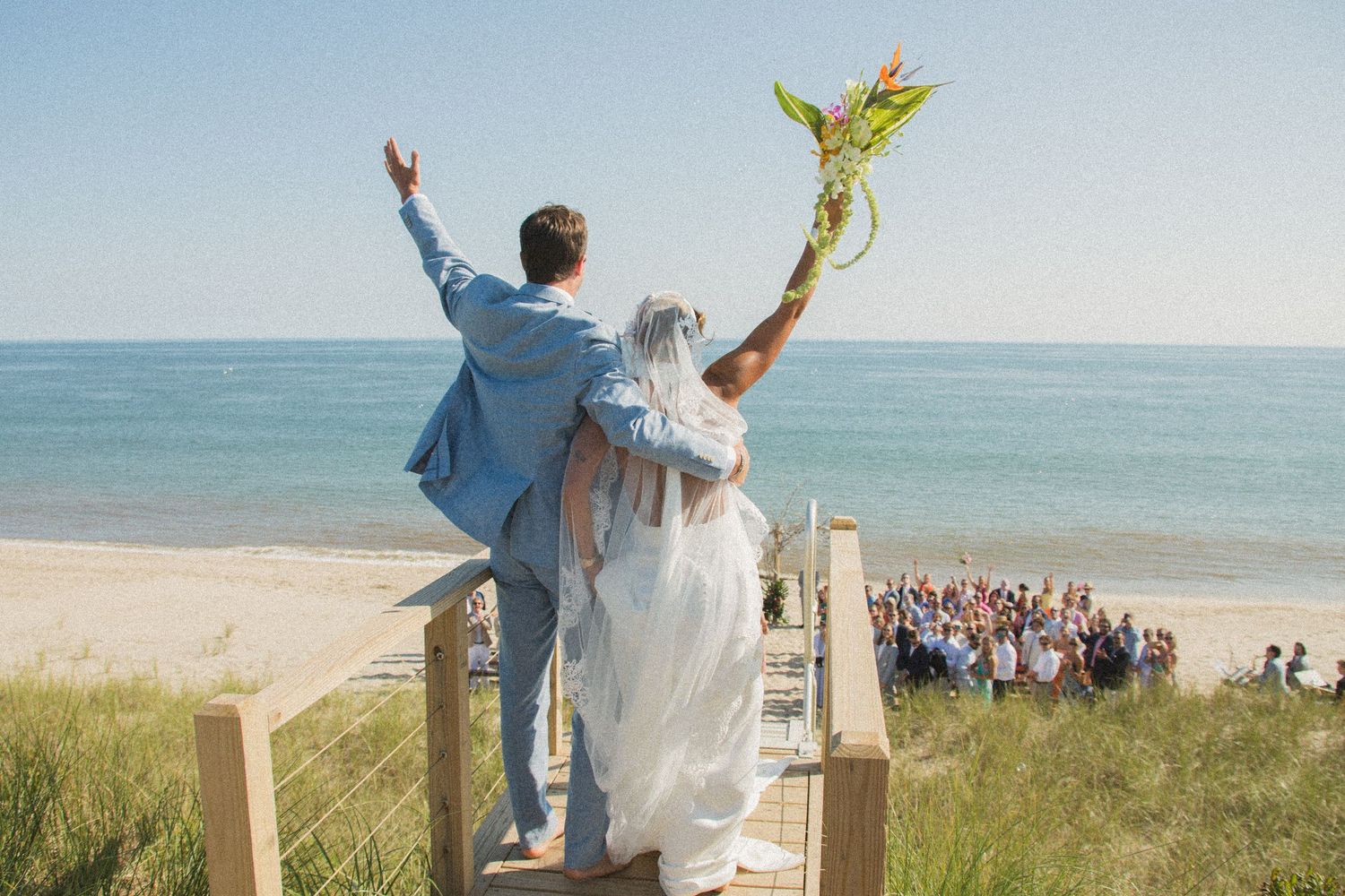Couple celebrates on beach boardwalk with guests cheering below on the sand.
