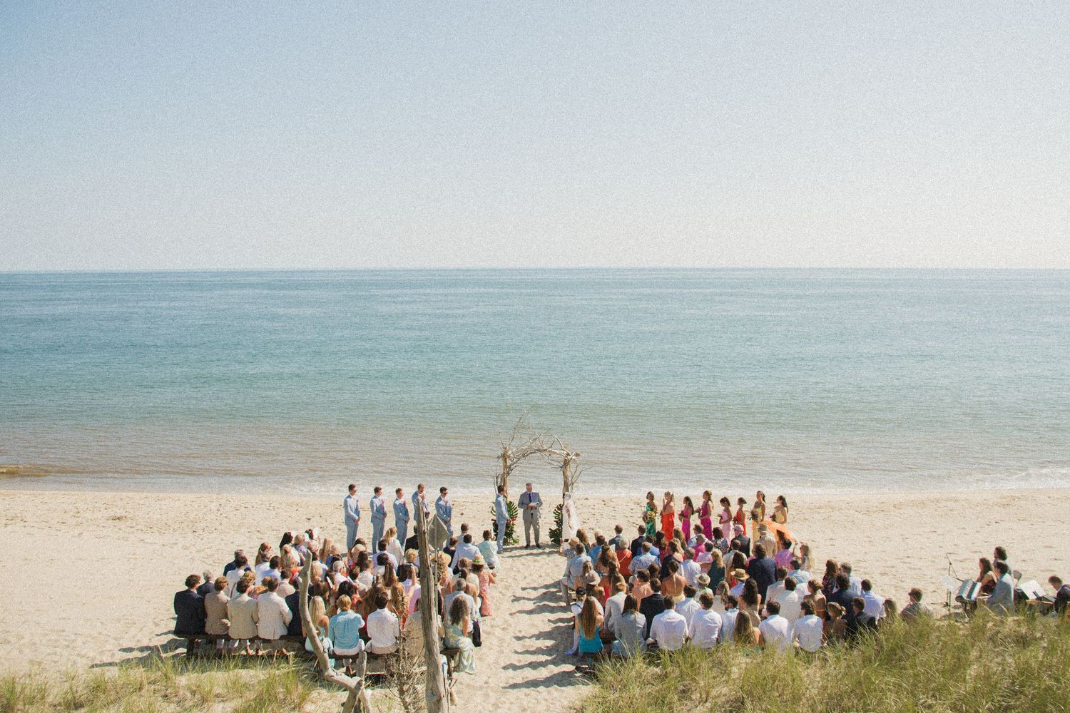 Panoramic view of beach wedding ceremony with guests gathered by the ocean.