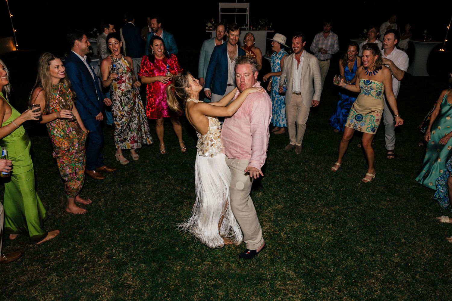 Large group of wedding guests dancing on a lawn under evening string lights.