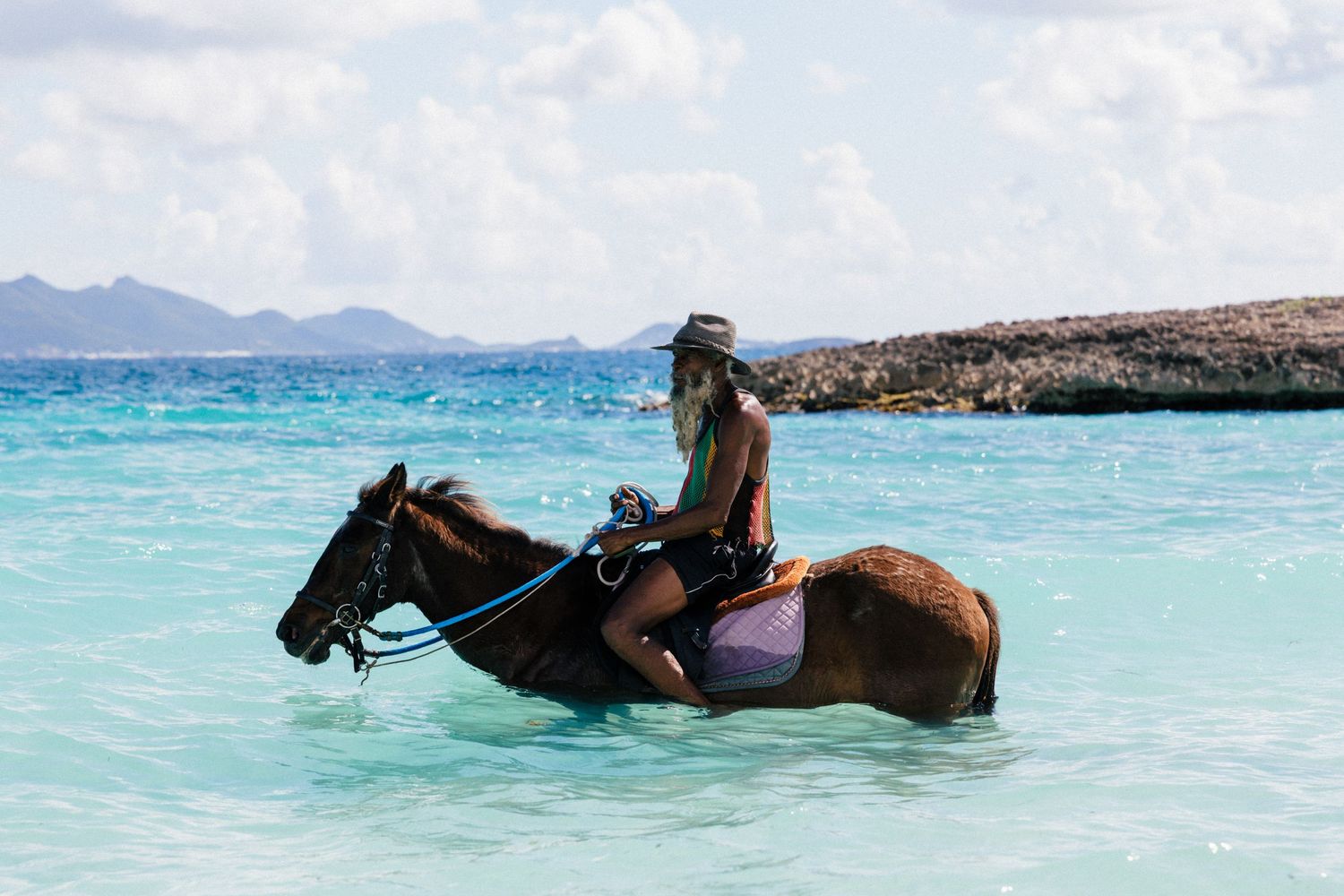 Horse riding on pristine beach with sailboat and mountains visible in distance.