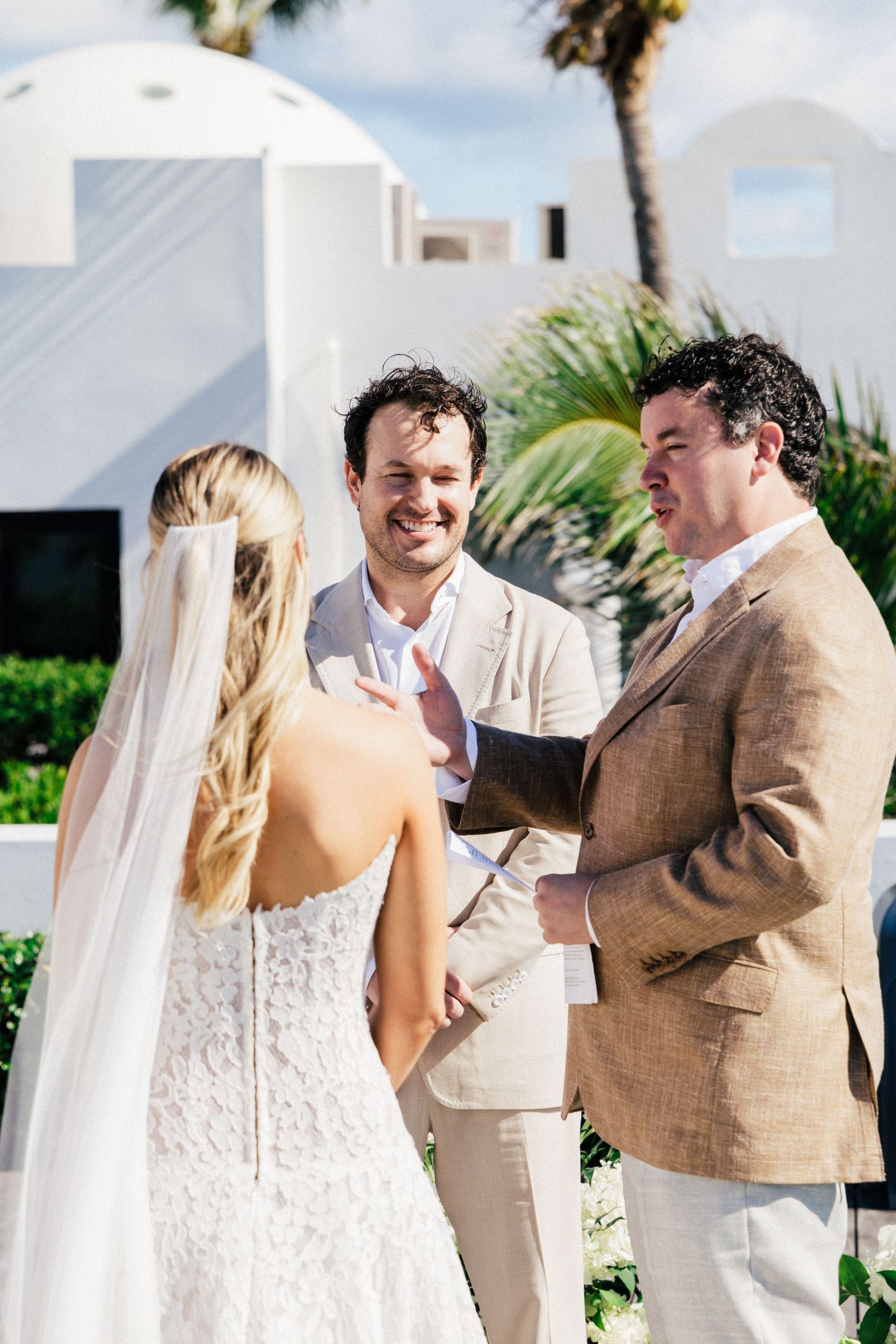 A beach wedding ceremony with the couple and officiant standing together in light-colored attire against an ocean backdrop.