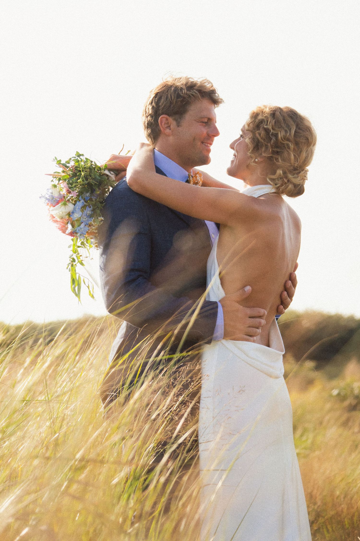 A couple embraces in a sunlit field of tall grass during their wedding celebration.