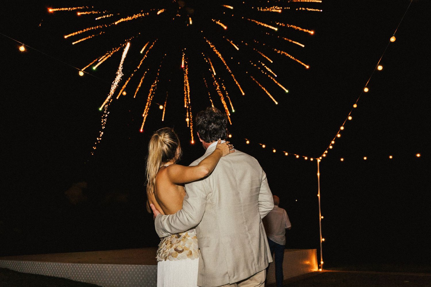 A couple embraces under fireworks during a nighttime celebration at an outdoor wedding venue.