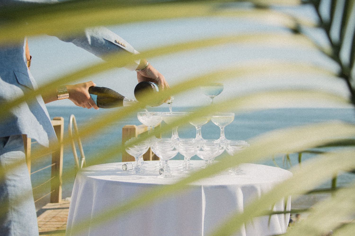 Champagne being poured into crystal glasses through palm fronds at beach event.