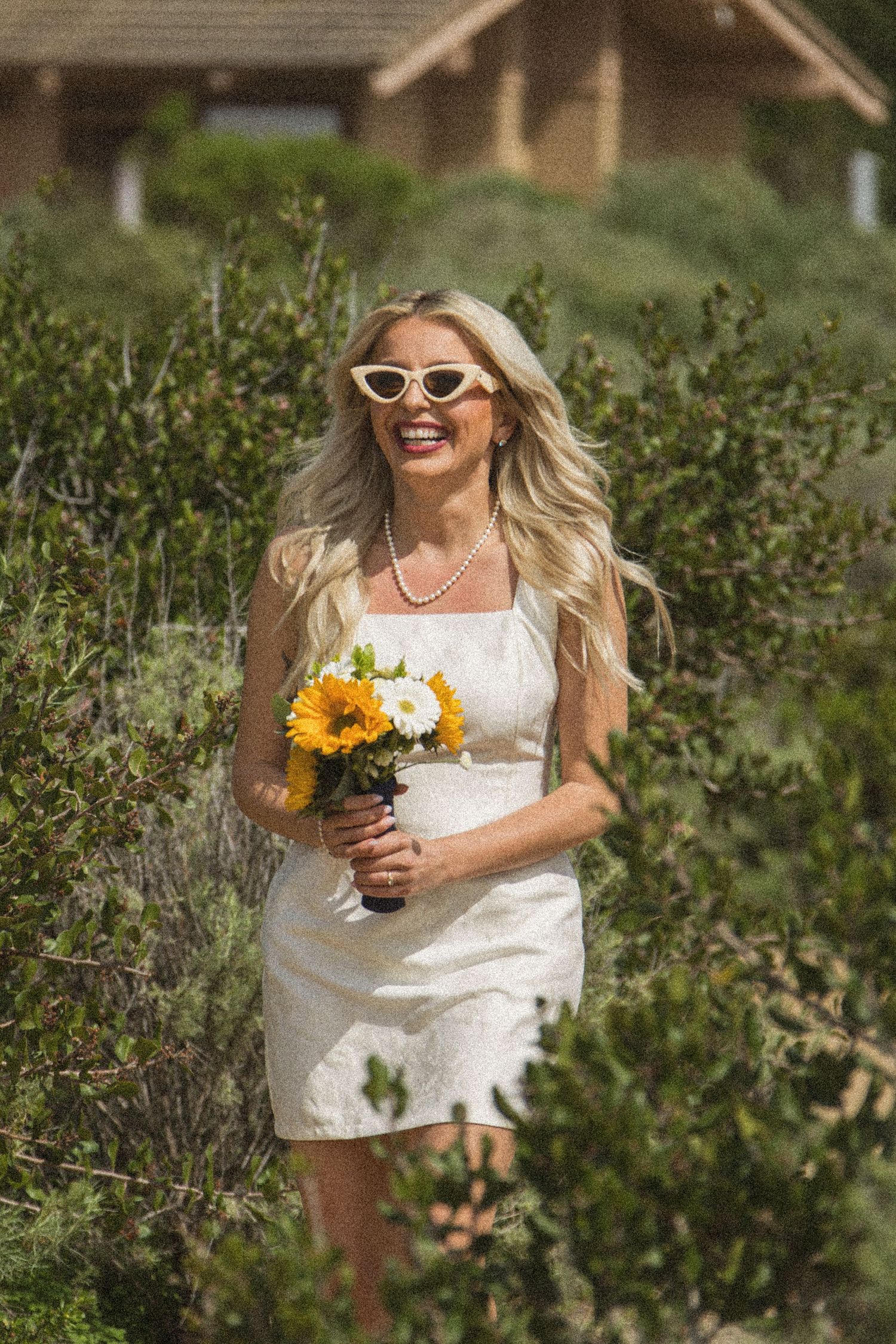 Bride in white dress holding sunflower bouquet standing among green shrubs.