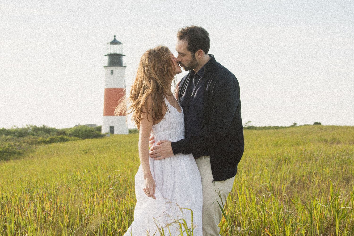 A romantic moment captured in a field with a lighthouse silhouetted against the sky.