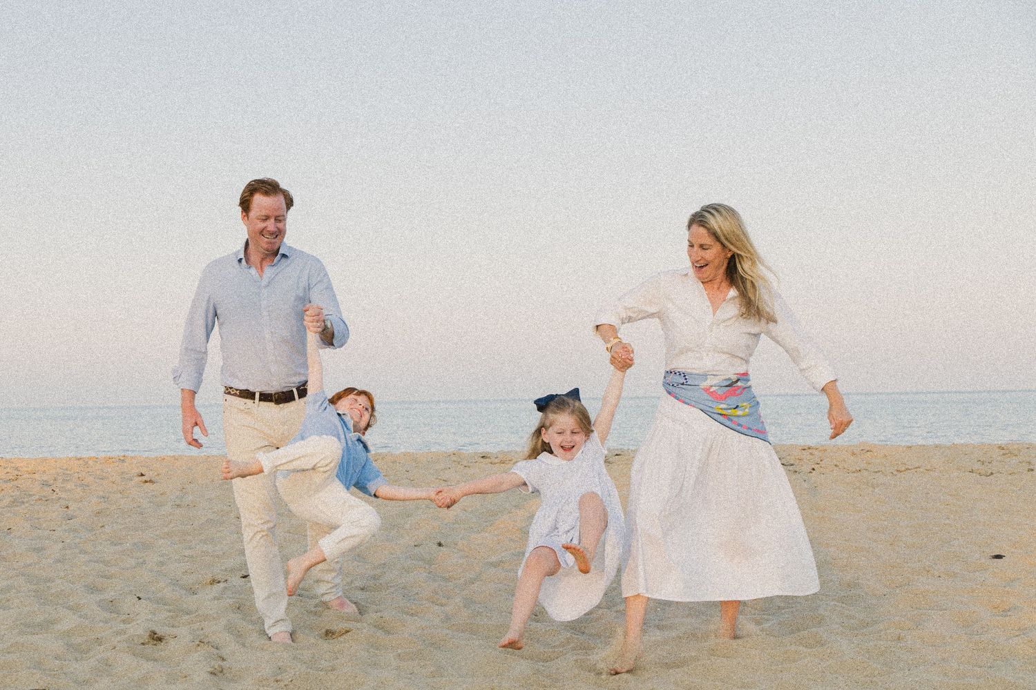 Family of four playing on a sandy beach, parents swinging children by hands, wearing white and blue outfits.