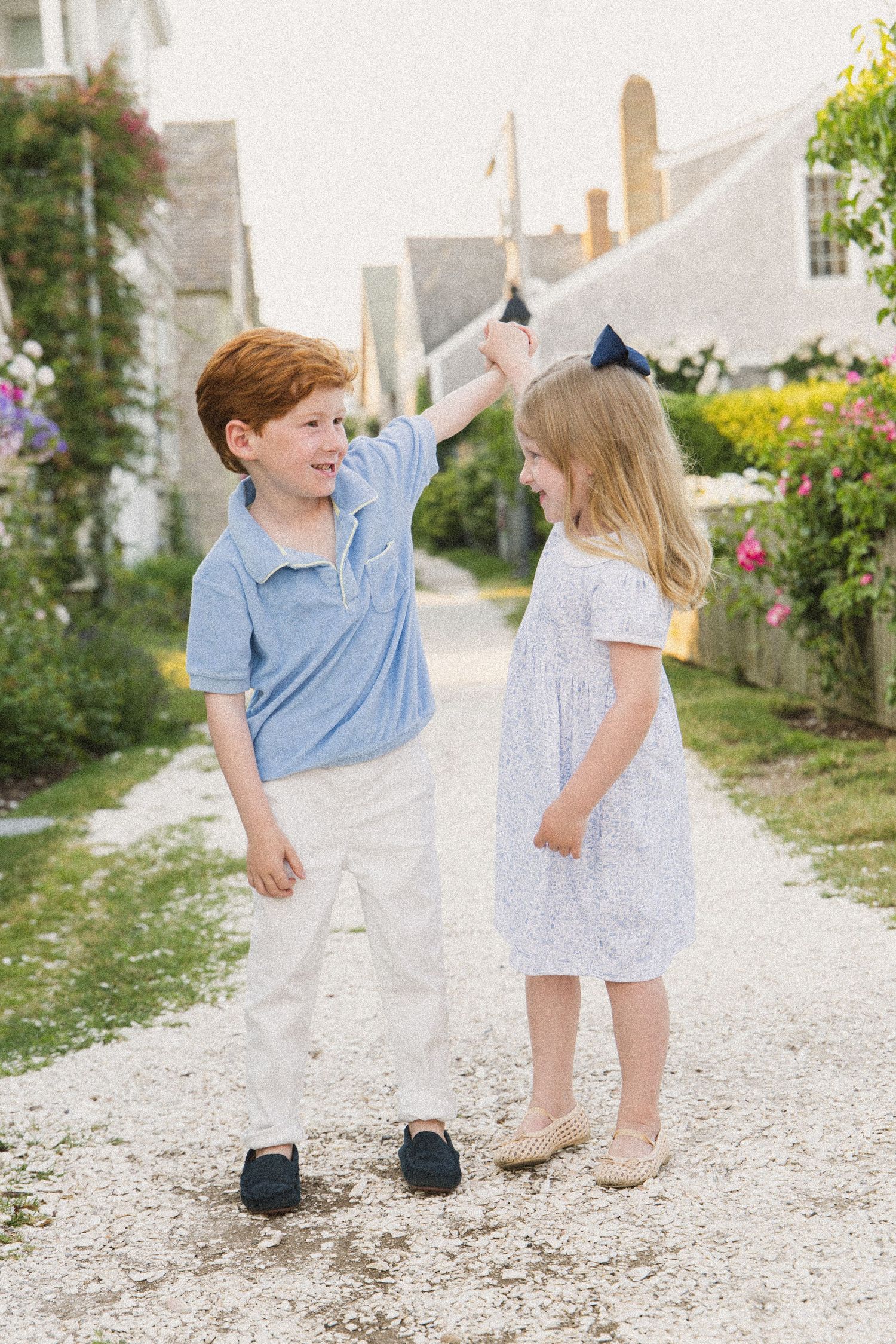 Two young children in preppy outfits hold hands on a gravel path surrounded by flowers and charming cottages.