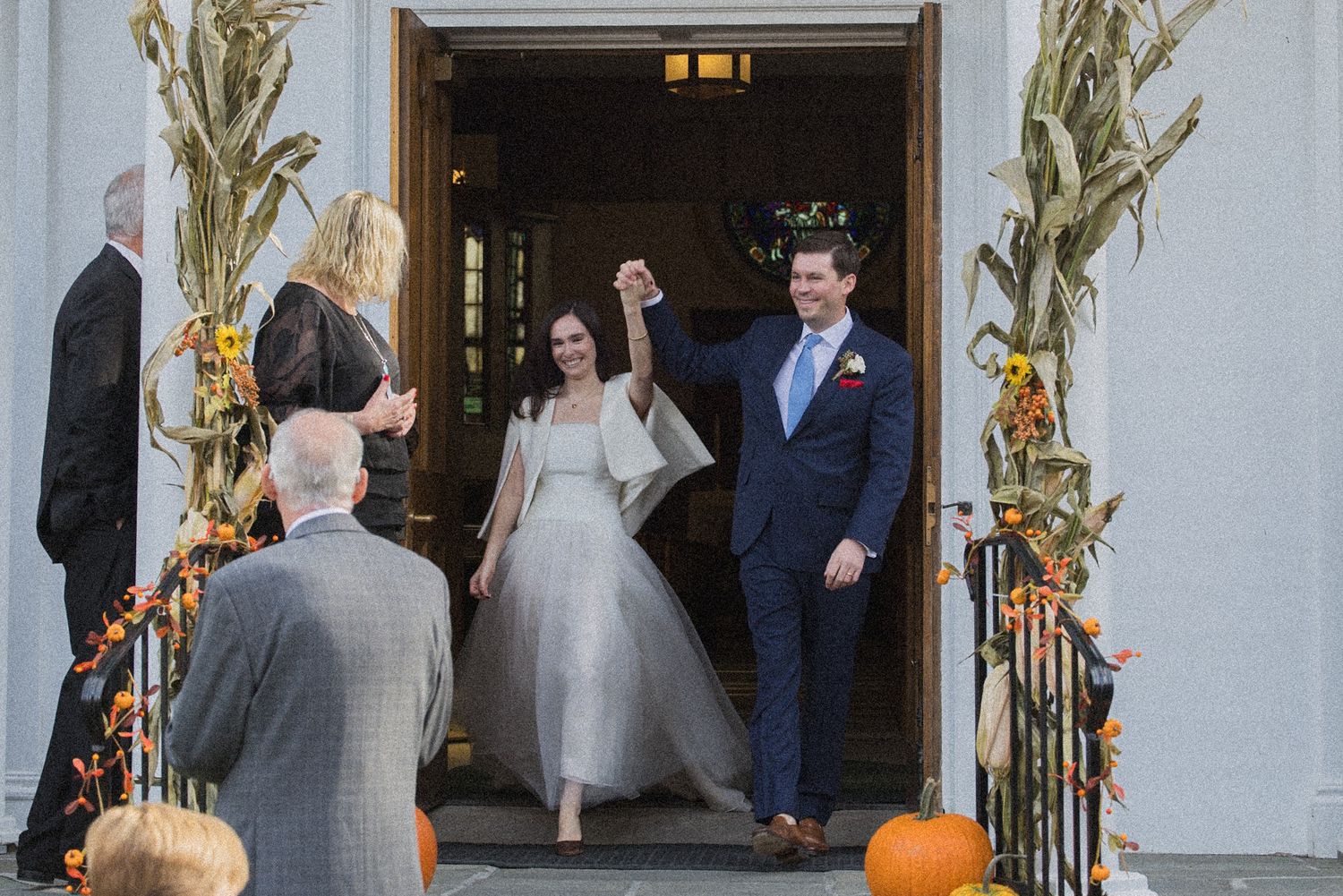 A bride in a white gown and a groom in a navy suit raise joined hands in a church doorway as guests watch beside autumn corn stalk decor.