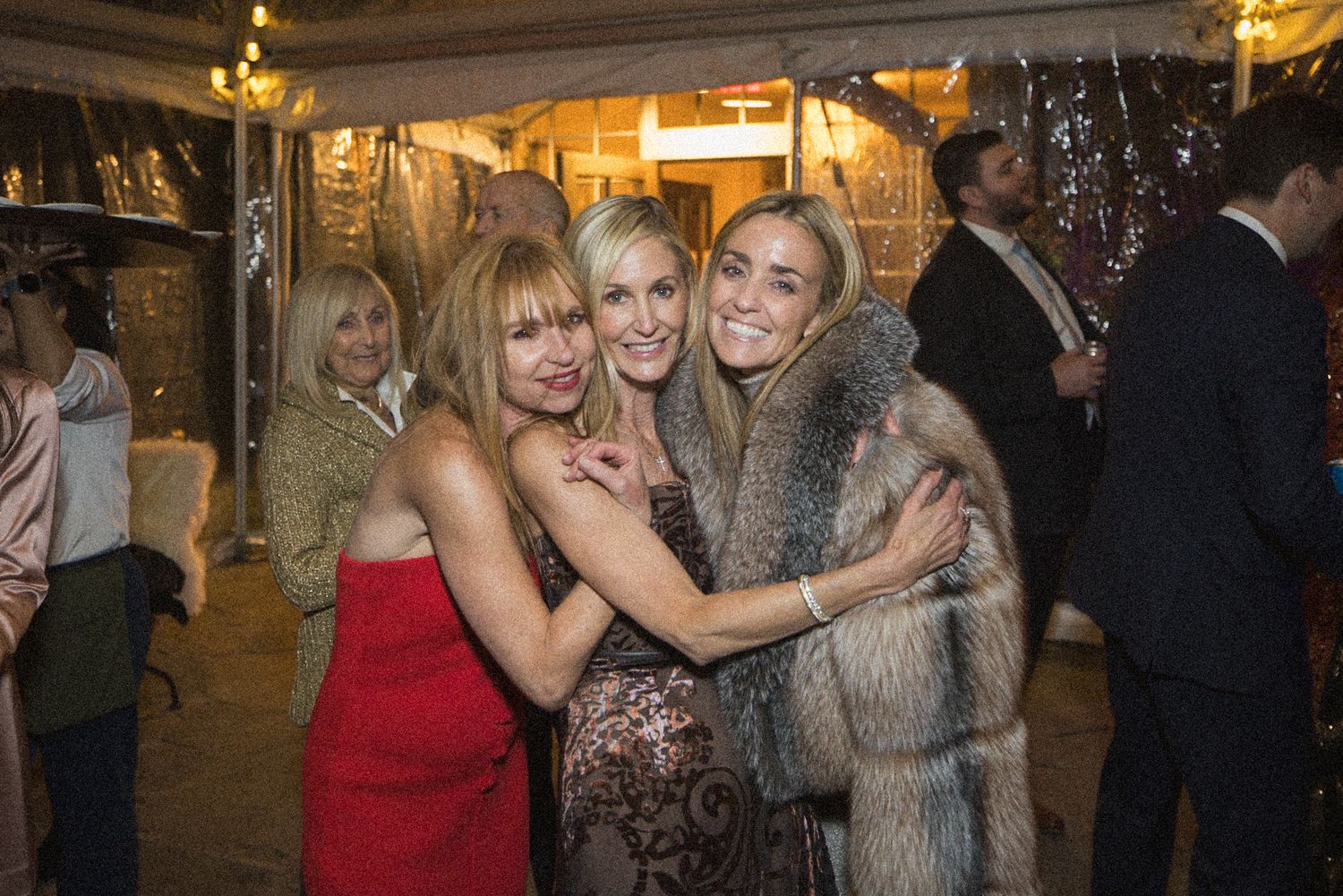 Three smiling women in elegant dresses and a fur coat embrace closely at a festive evening party with guests mingling in the background.