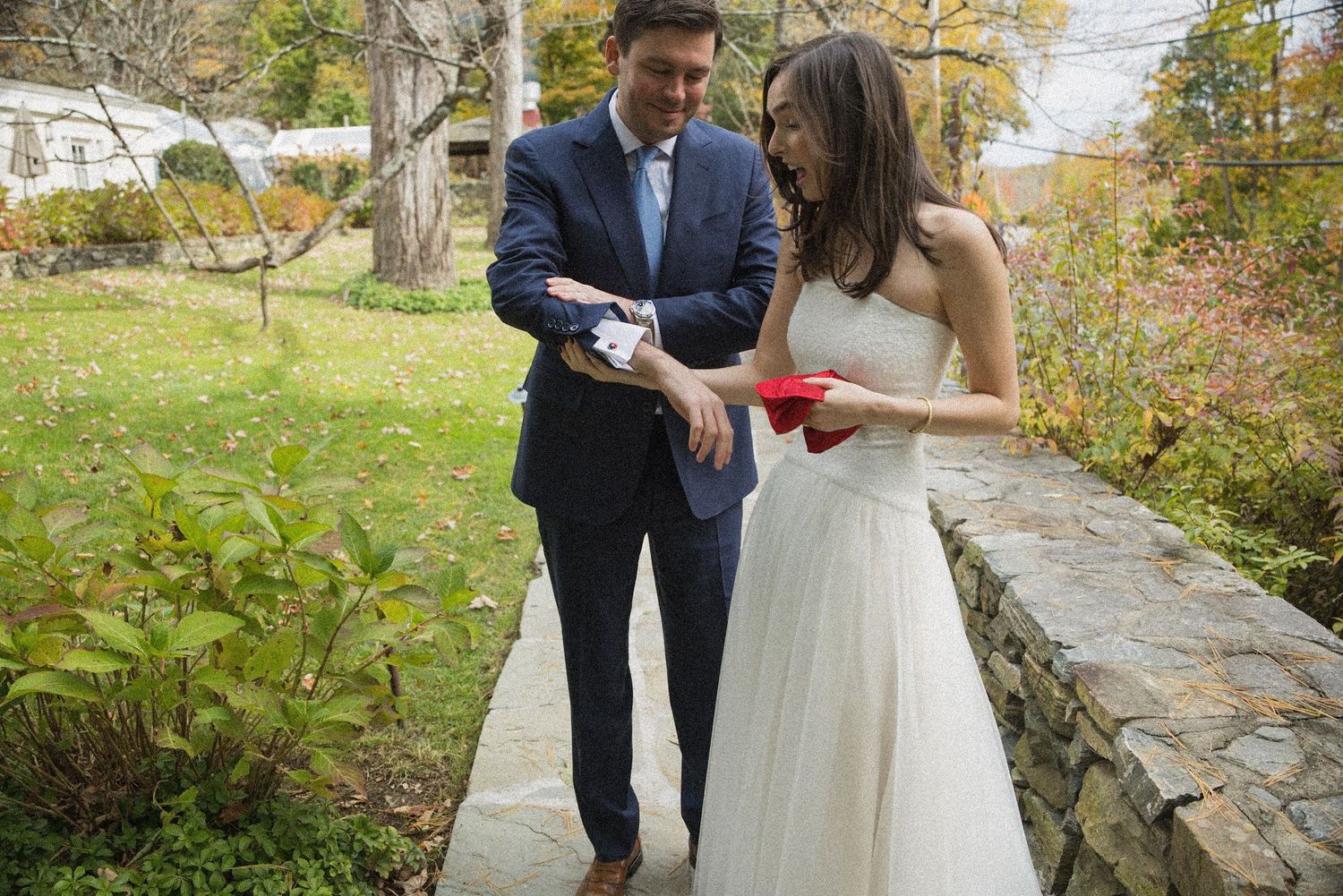 A groom in a navy suit and a bride in a white wedding gown stand on a garden path, looking at a small red item in her hands.