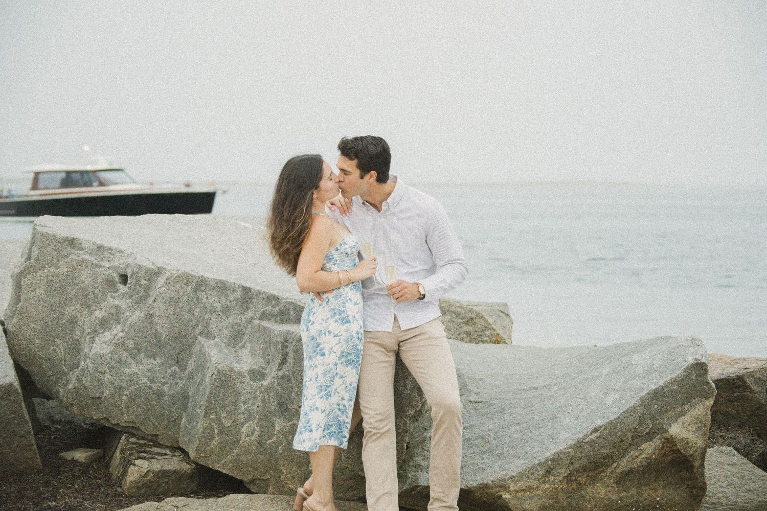 Couple sharing a romantic kiss near rocky shoreline with calm ocean waters in the background.