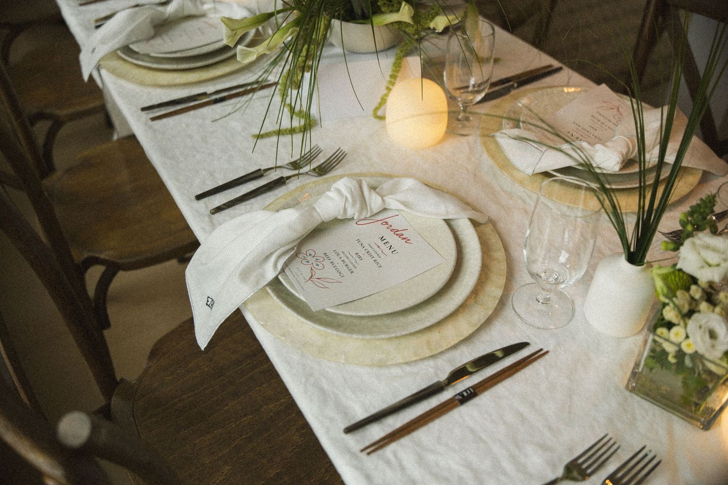 Elegant wedding table setting with white linen, layered plates, crystal glassware, candles, and fresh greenery centerpieces.