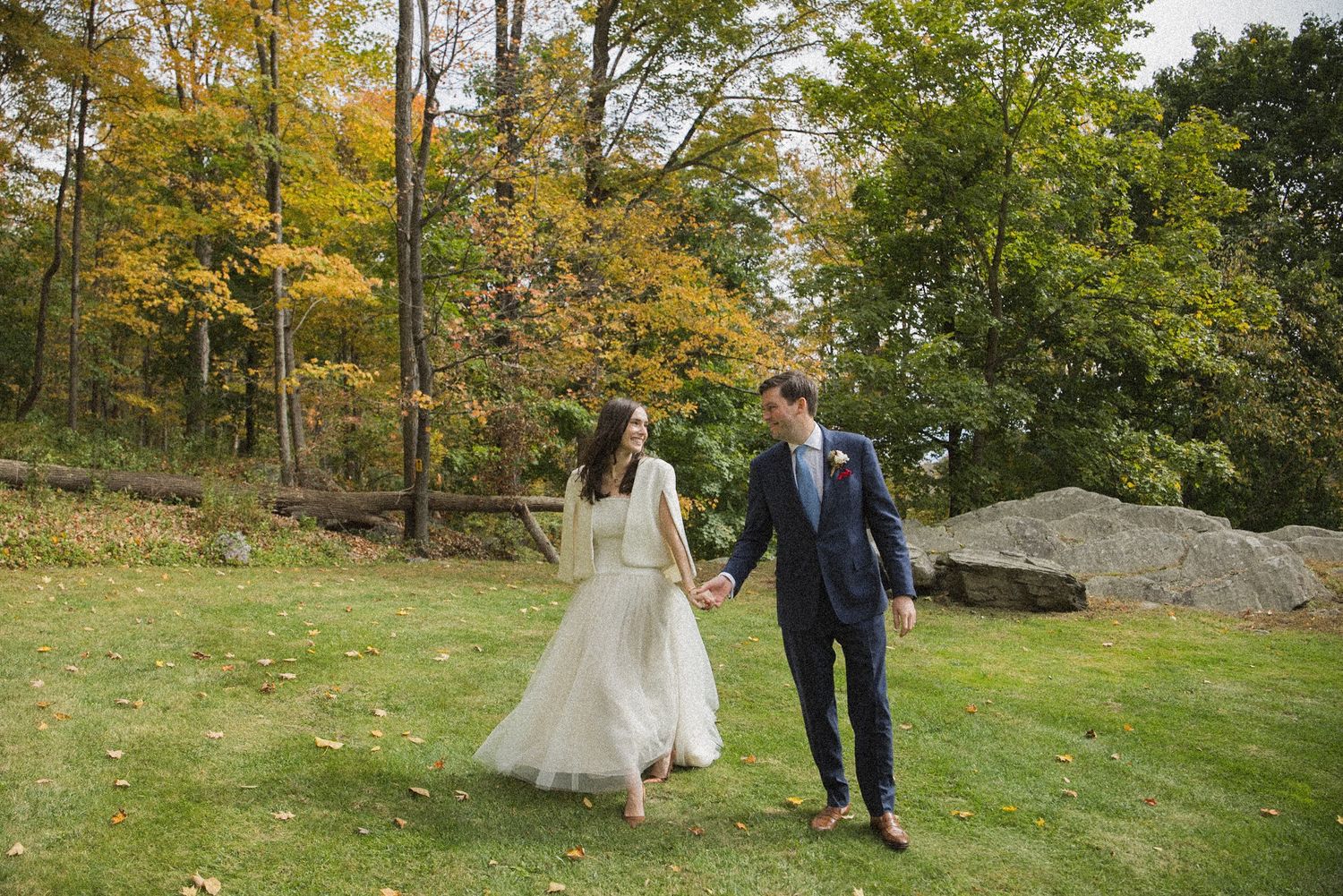 Bride in a white gown and groom in a navy suit hold hands while walking across a grassy lawn framed by autumn trees.