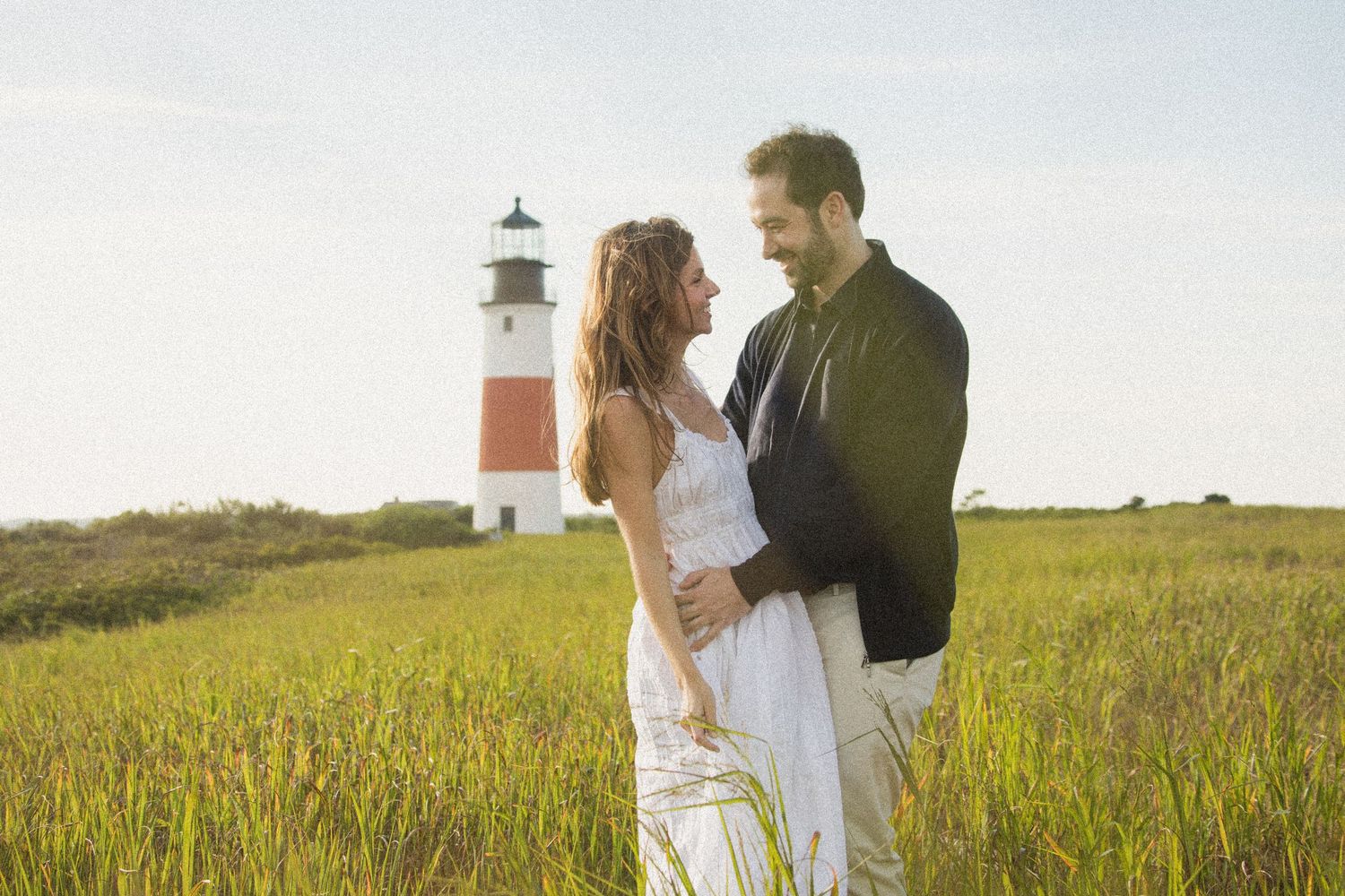 Couple walking through tall grass field with lighthouse in background during sunset.