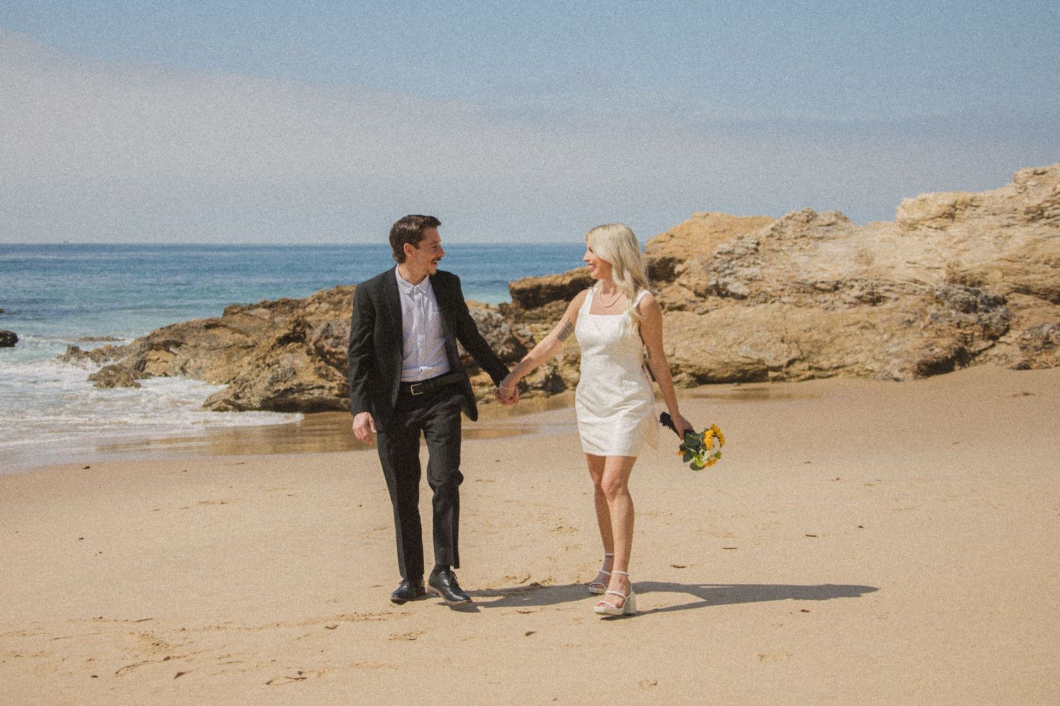 Wedding couple walks along a sandy beach with coastal cliffs and ocean views.