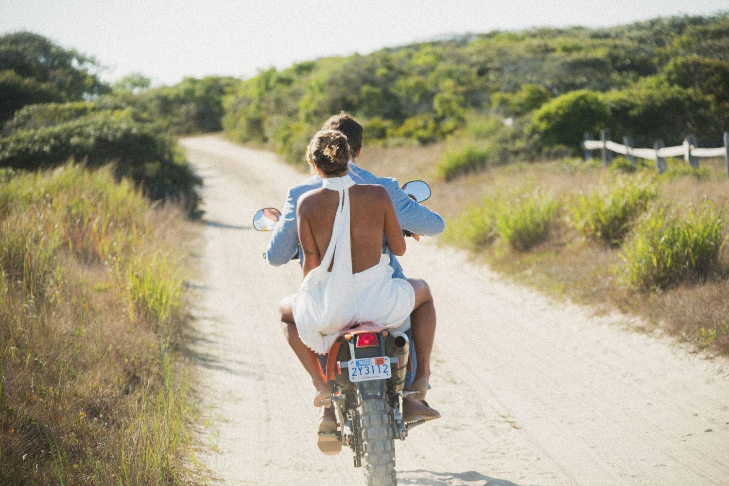 Back view of newlyweds riding away on motorcycle down sandy beach path.