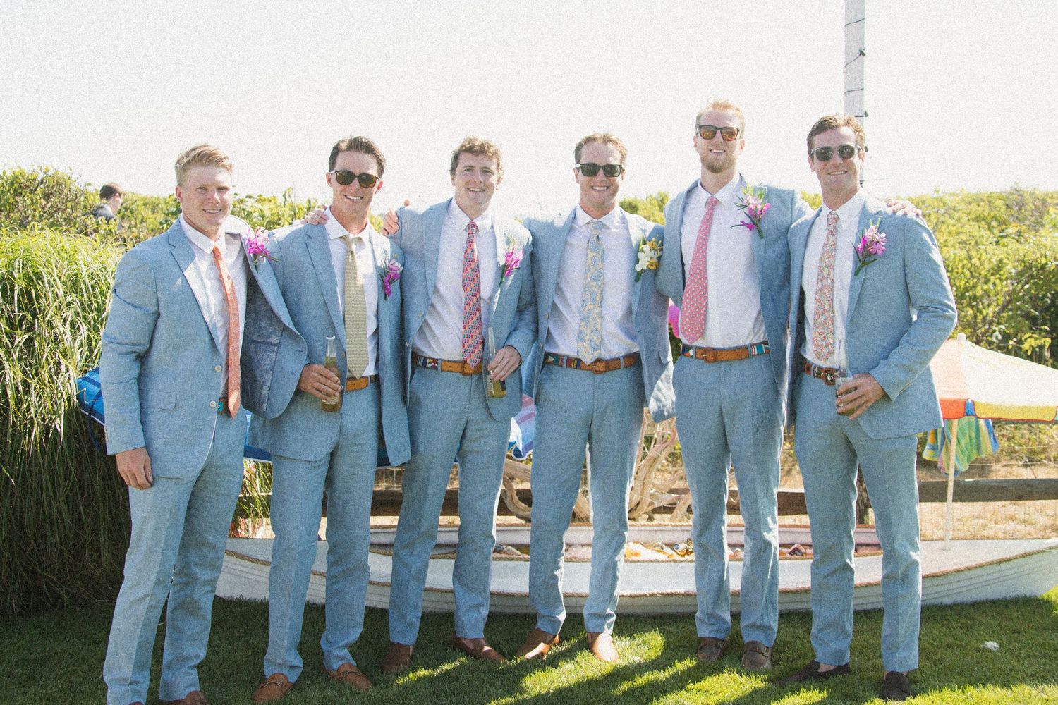 Groomsmen in light blue suits with coral ties pose together outdoors on a sunny day.