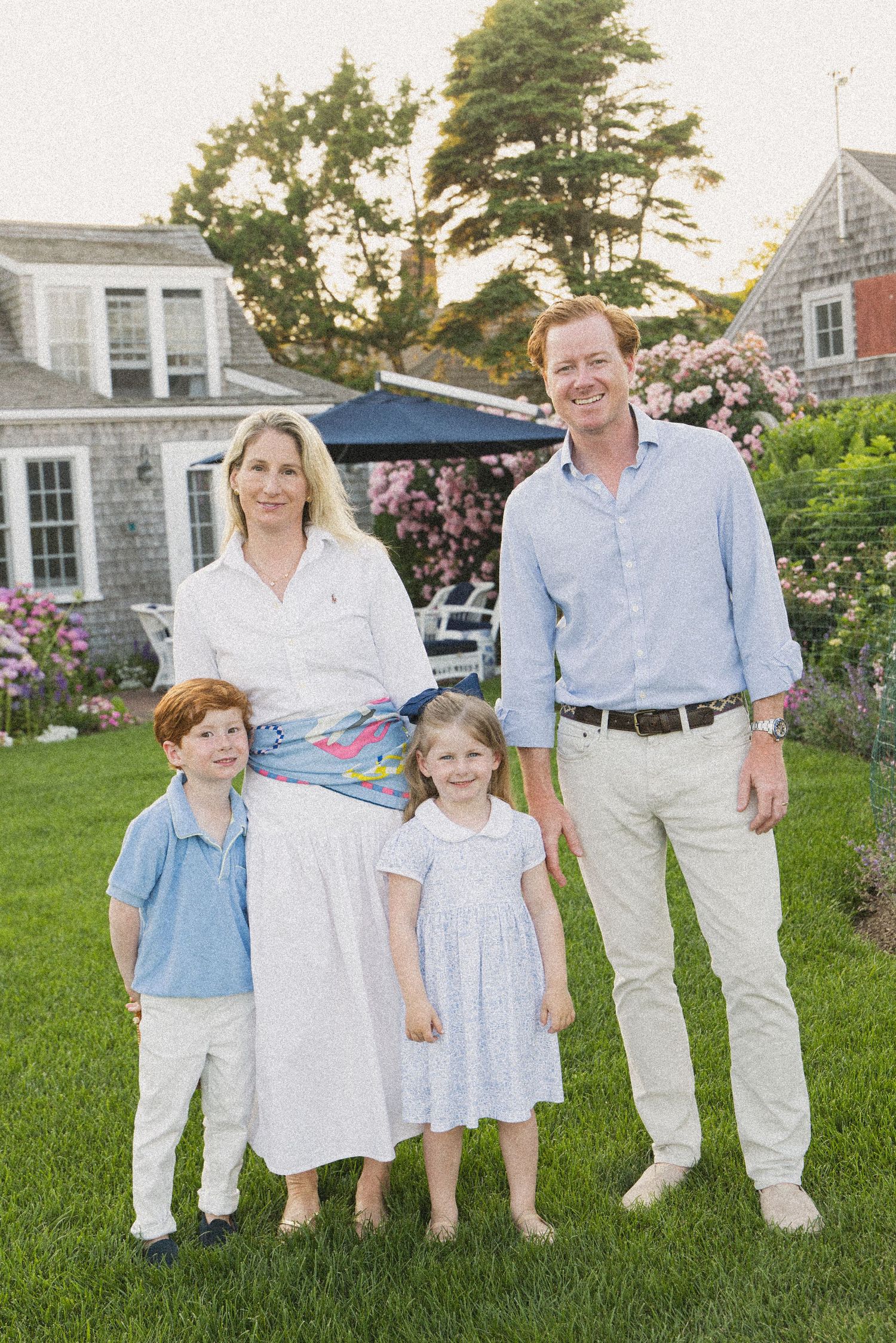 Family of four posing in a lush green backyard near a charming cottage with blooming flowers.