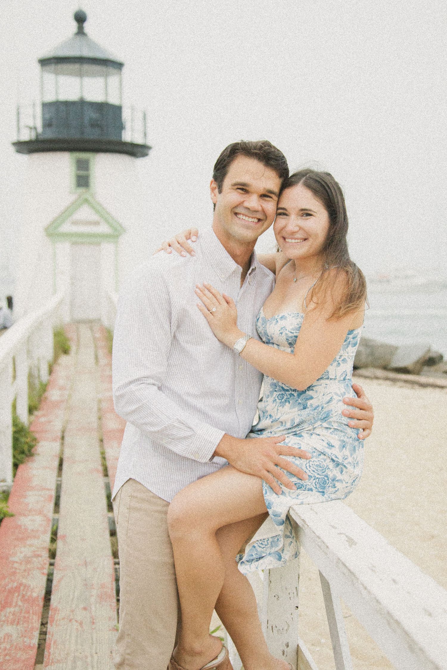 Couple embracing and smiling near a lighthouse on a sunny day, woman in floral dress, man in white shirt.