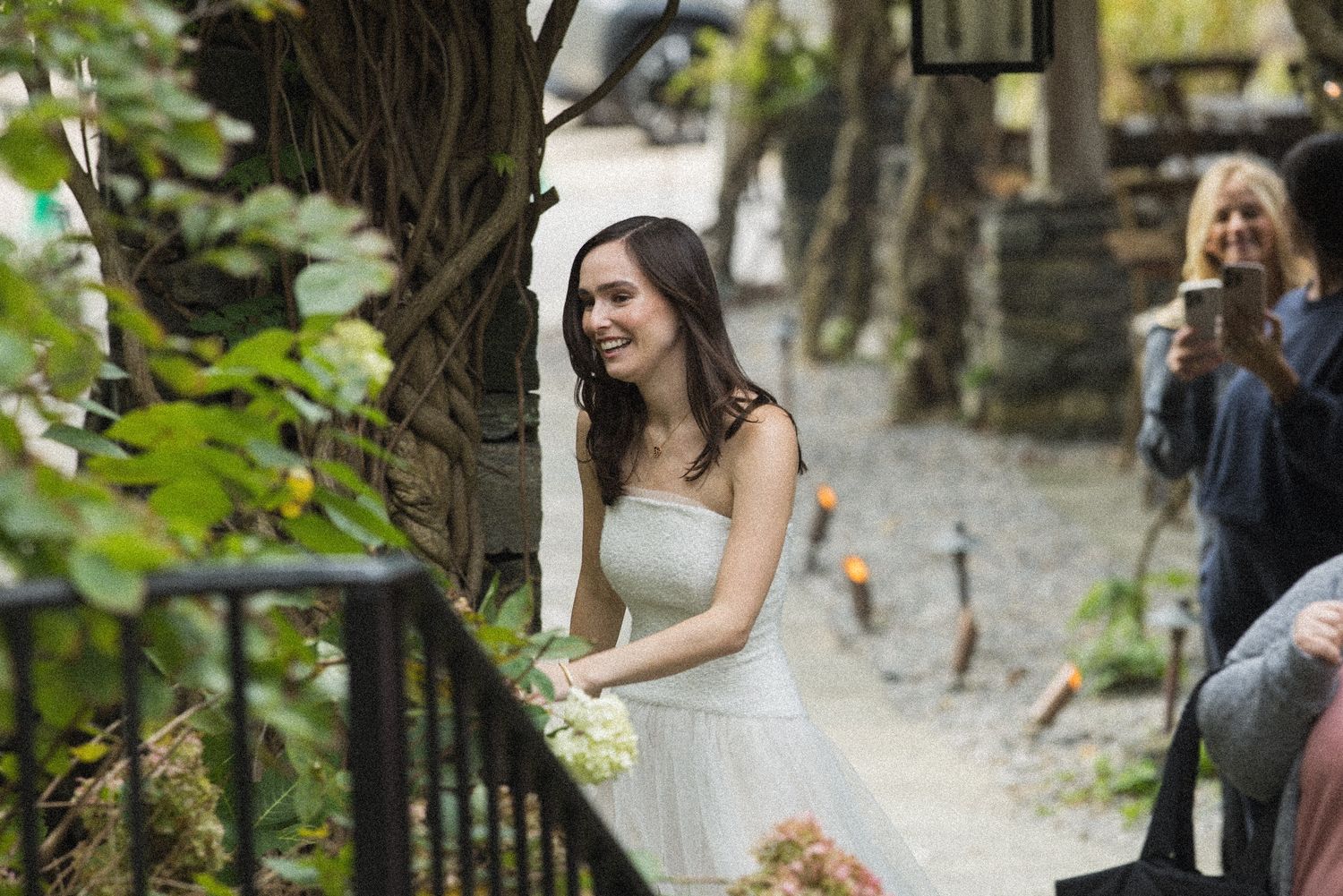 Smiling bride in a white strapless wedding dress holds a bouquet while walking along a garden path as guests photograph her.