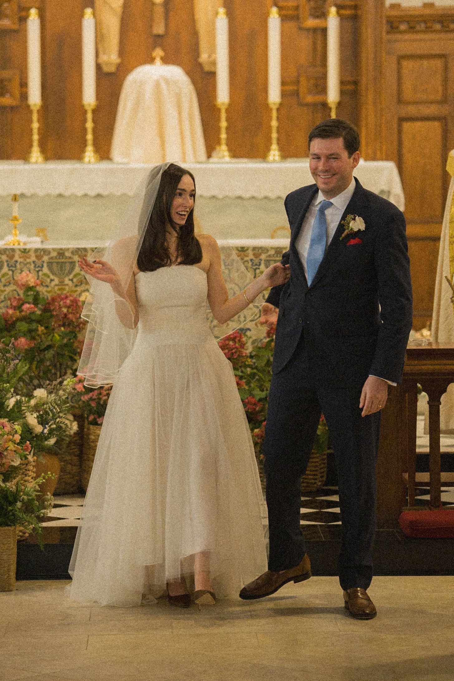 Smiling bride in a strapless tulle wedding gown and veil holds hands with a groom in a navy suit and blue tie inside a church.